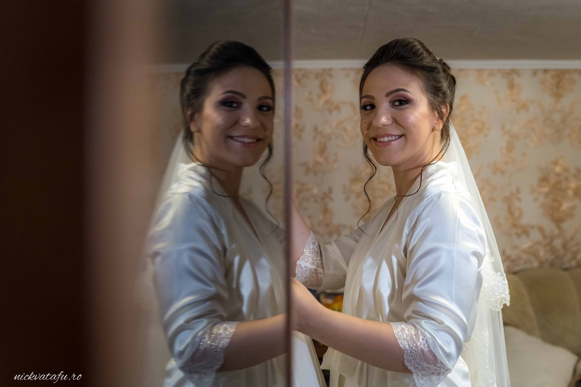 Mireasă zâmbitoare în rochie albă, pregătindu-se pentru nuntă, reflecție emoționantă în oglindă.