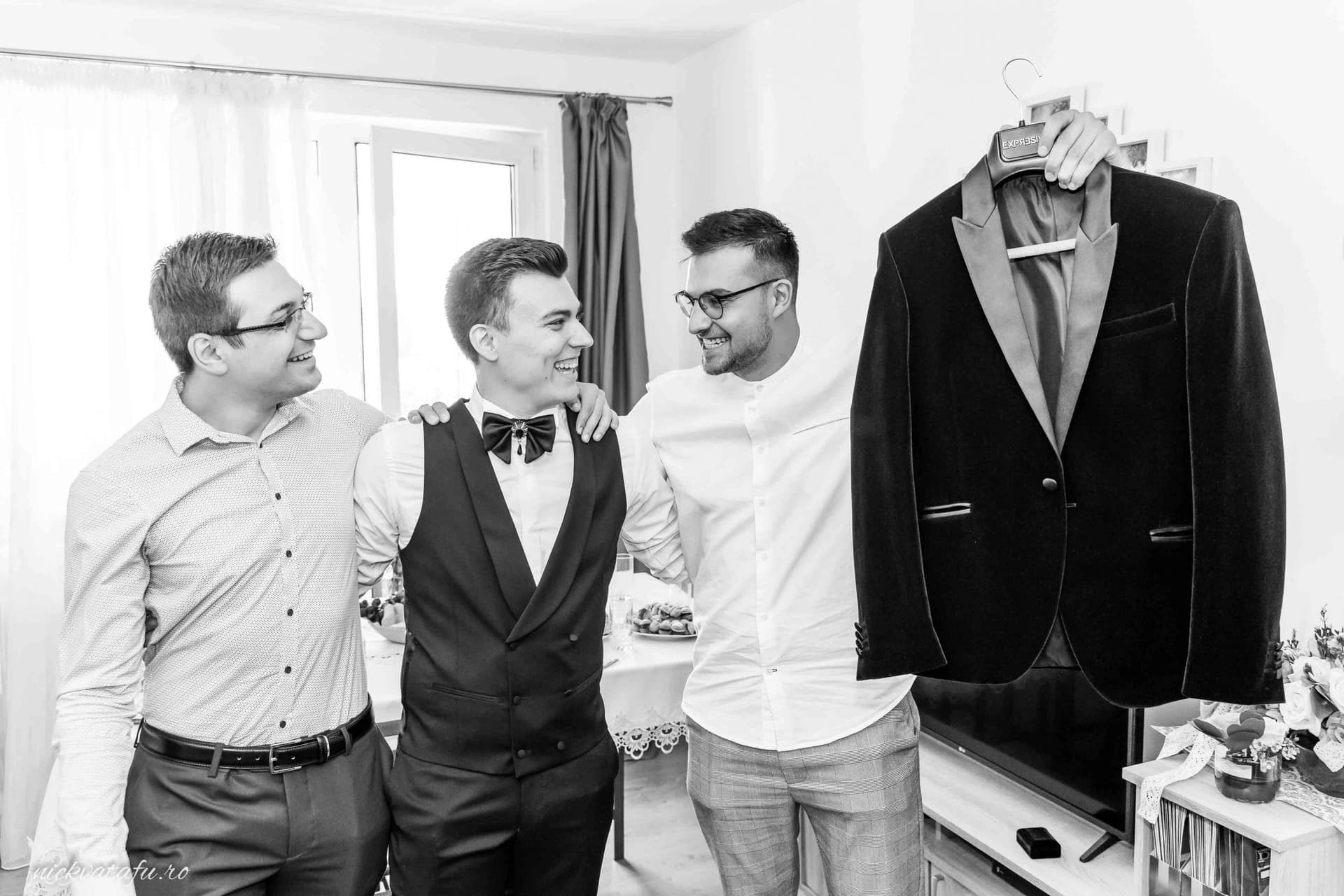 Prietenii mirelui se pregătesc cu emoție înainte de nuntă, în atmosfera intimă a unei camere luminoase.