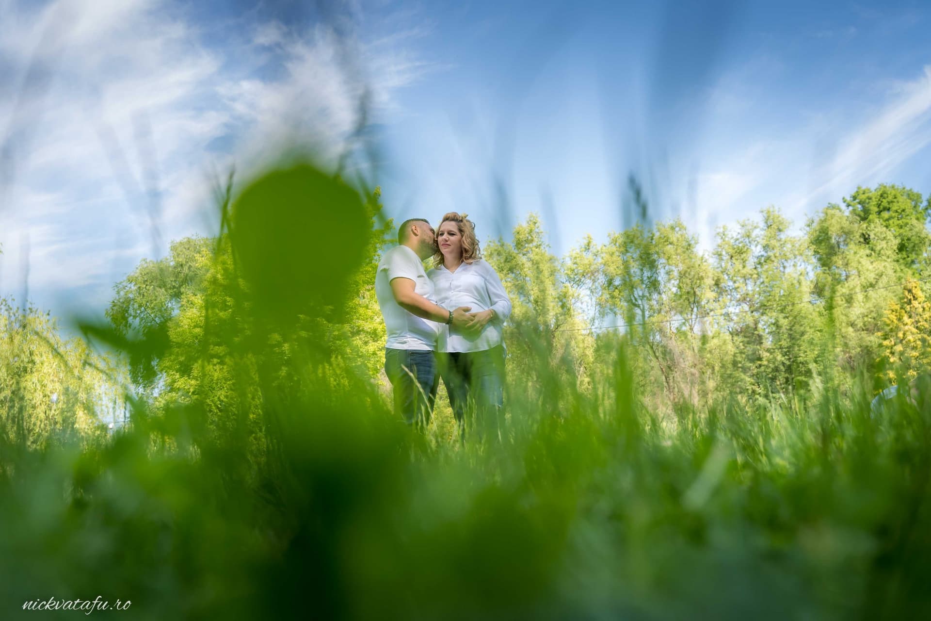 Îmbrățișare plină de emoție între doi îndrăgostiți, fotografie de nuntă realizată în natură, cu iarbă verde în prim-plan.