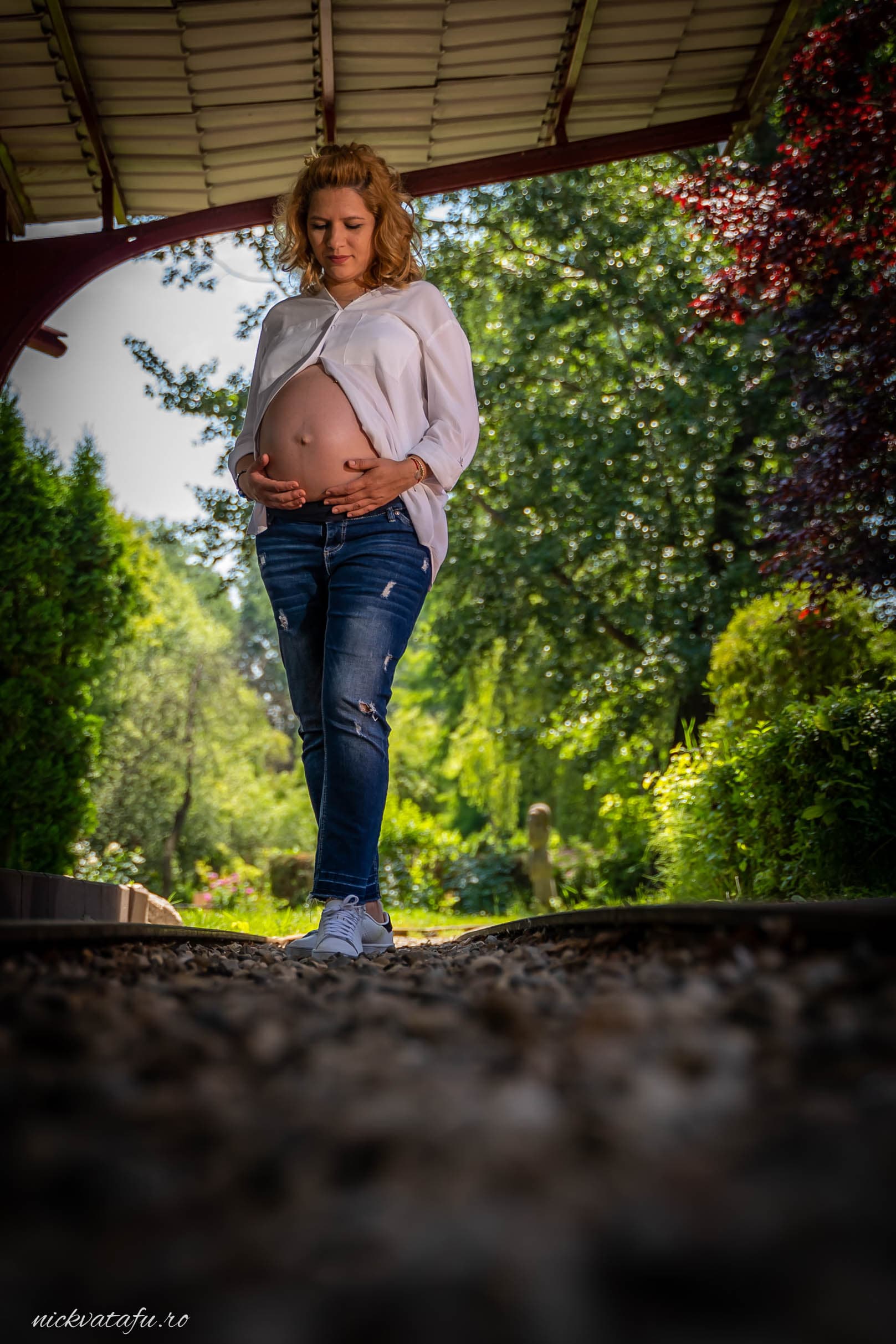 Femeie însărcinată, zâmbitoare, admirând natura într-o grădină verde, fotografie de maternitate plină de emoție.
