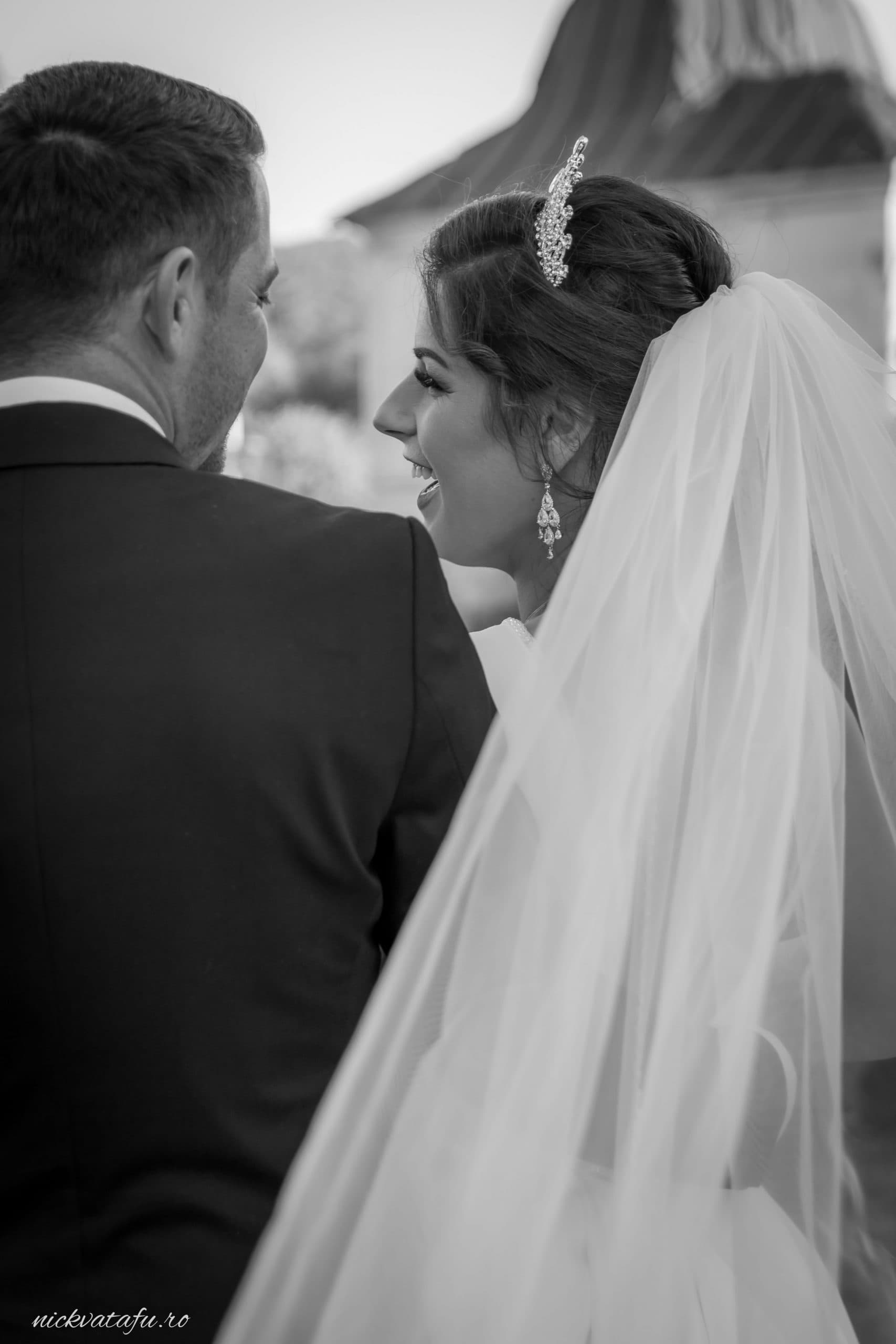 Zâmbete pline de emoție între mire și mireasă, în fotografie de nuntă, cu voal elegant în fundal.