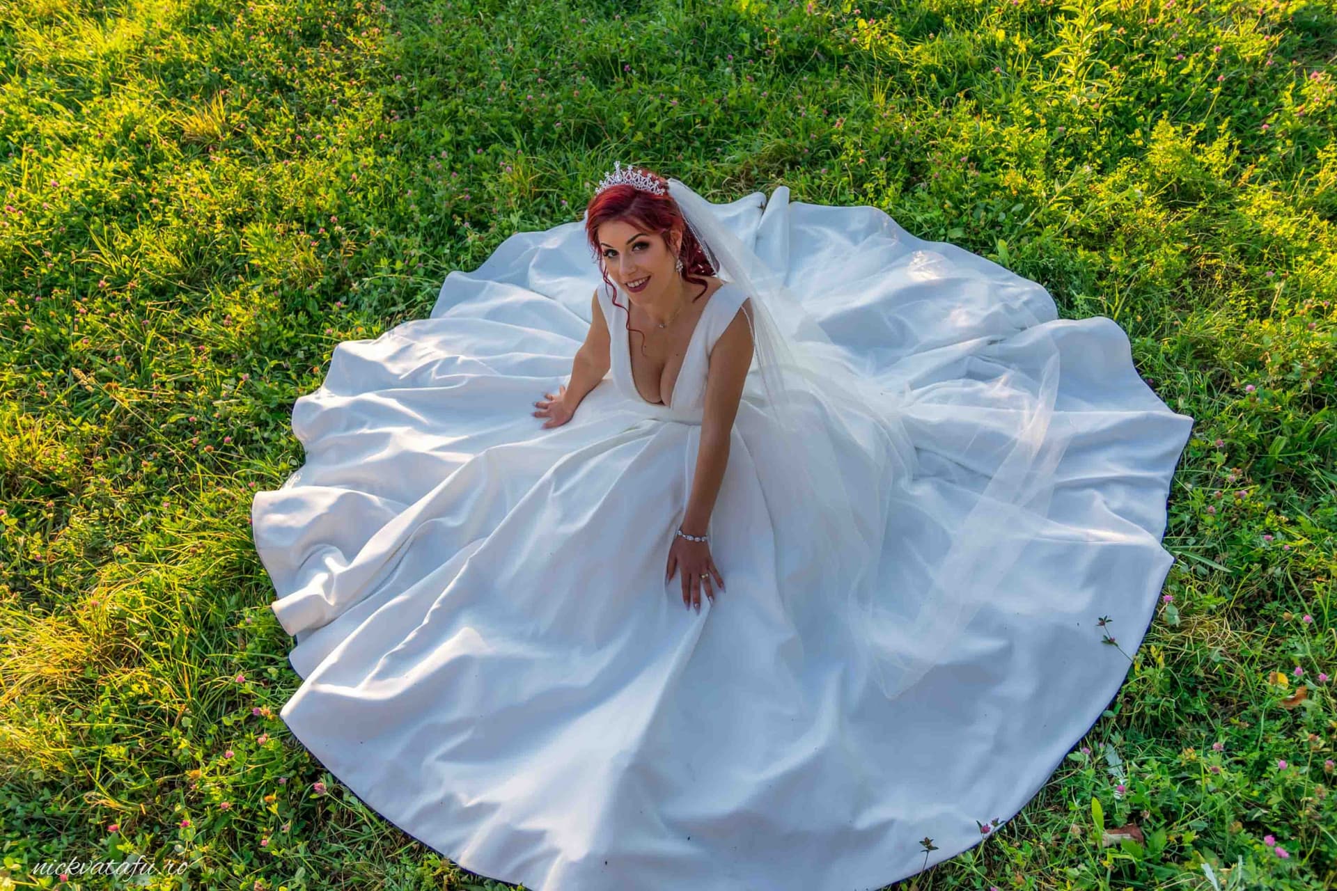 Mireasă zâmbitoare în rochie albă, așezată pe iarbă verde, capturând emoția unei fotografii de nuntă.