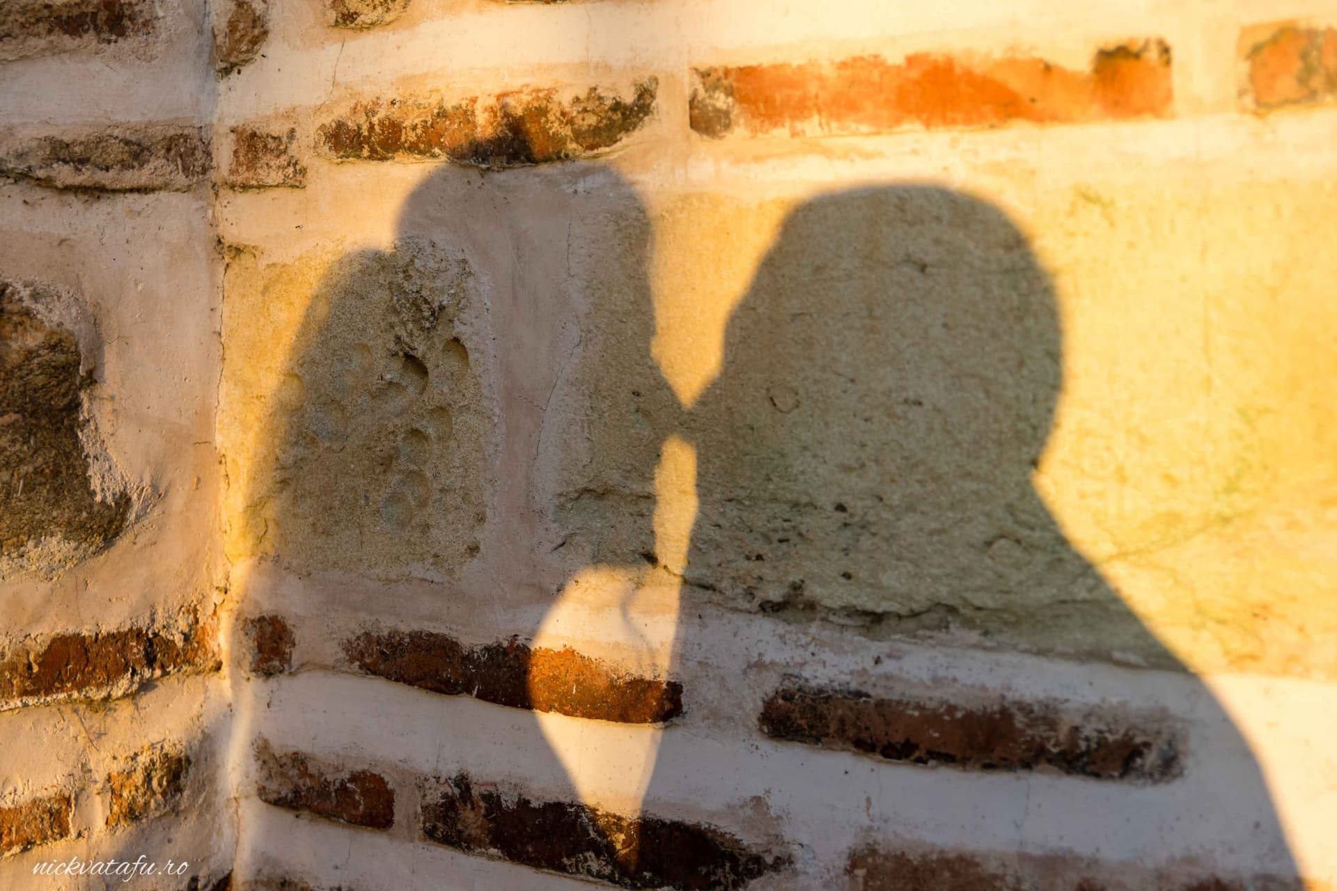 Siluetele a două persoane care se sărută, create de lumina soarelui pe un zid de cărămidă, evocând emoție și intimitate.