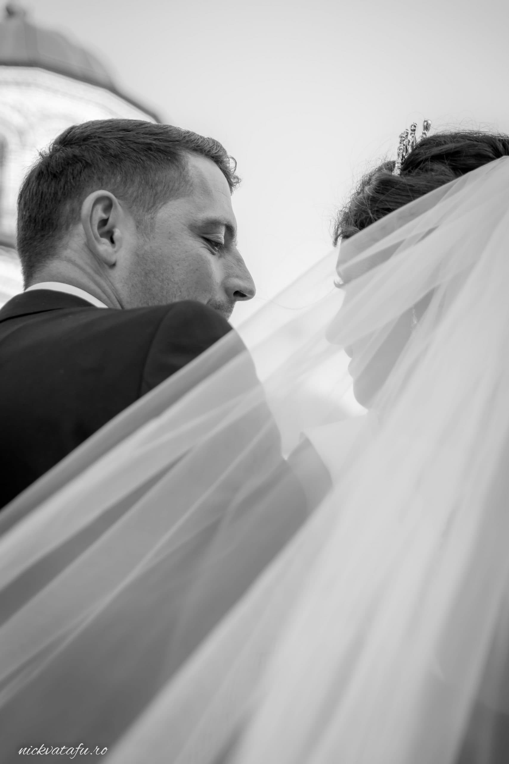 Mire și mireasă îmbrățișați sub voalul delicat, evocând emoție și romantism în fotografia de nuntă.