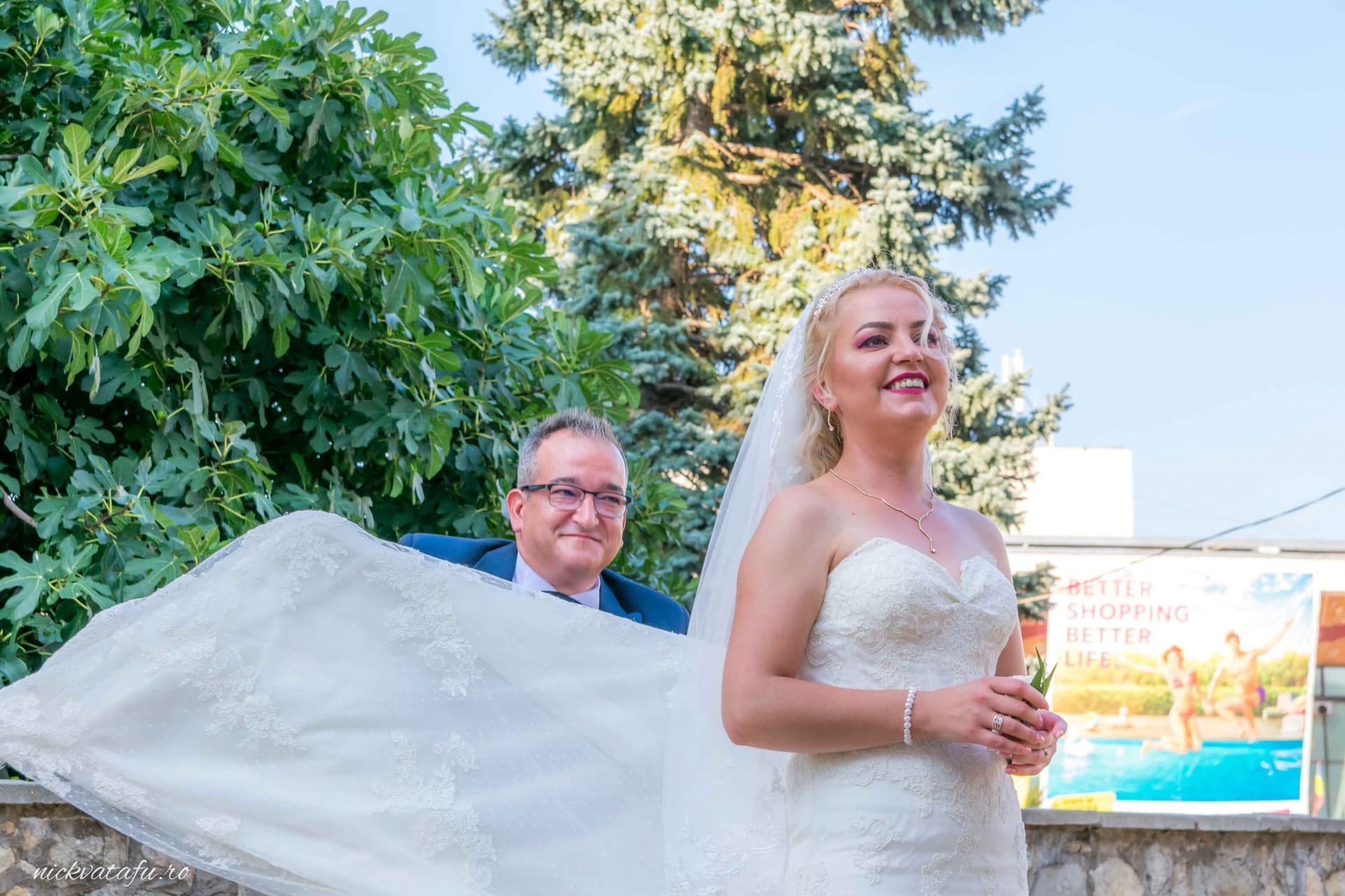 Mireasă zâmbitoare cu voal, însoțită de soț, în decor natural, fotografie de nuntă plină de emoție.