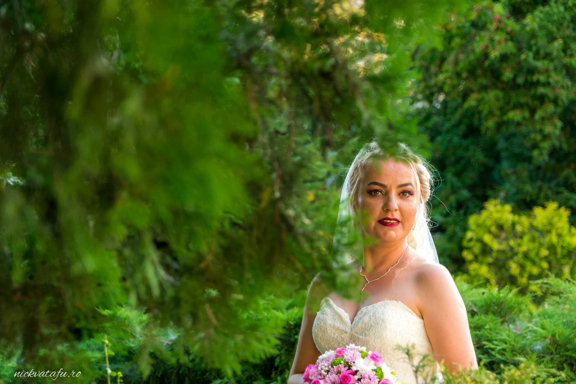 Mireasă zâmbitoare, cu buchet de flori, înconjurată de verdeață, fotografie de nuntă plină de emoție.
