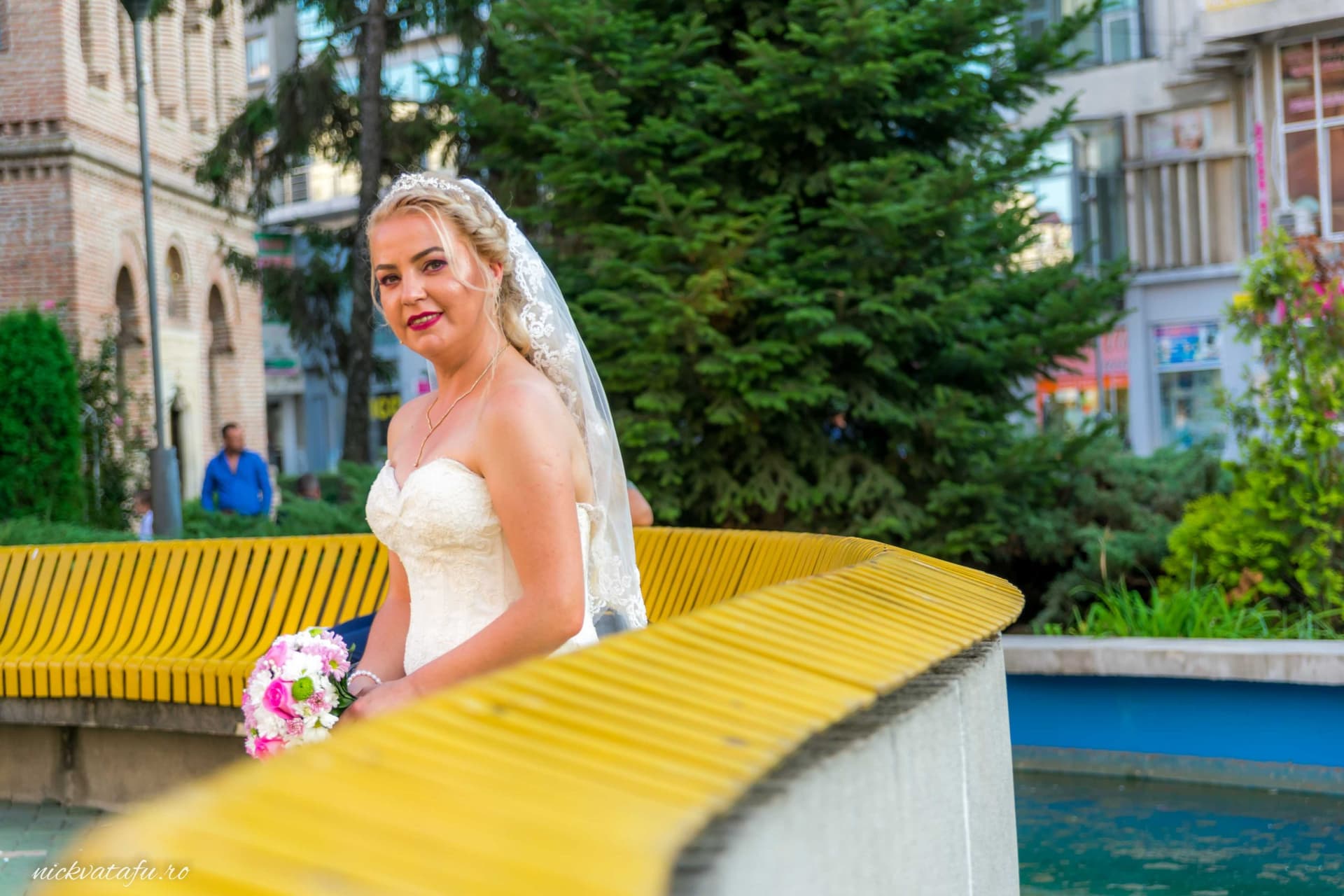 Mireasă zâmbitoare, în rochie albă, stând pe o bancă galbenă, capturând emoția unei zile de nuntă.