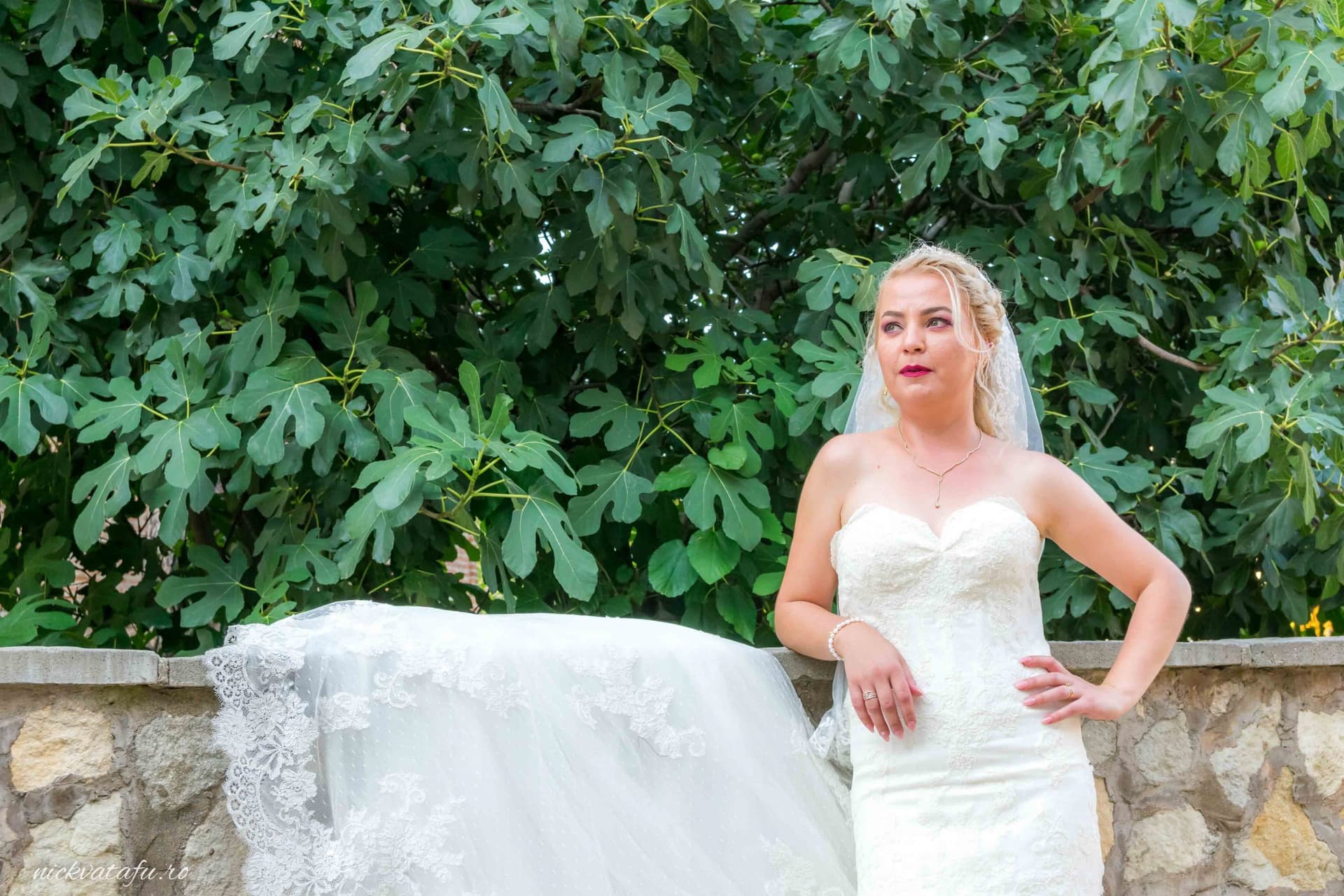 Mireasă elegantă, într-o rochie albă, pozând cu un zâmbet subtil printre frunzele verzi, detalii nuntă armonioase.