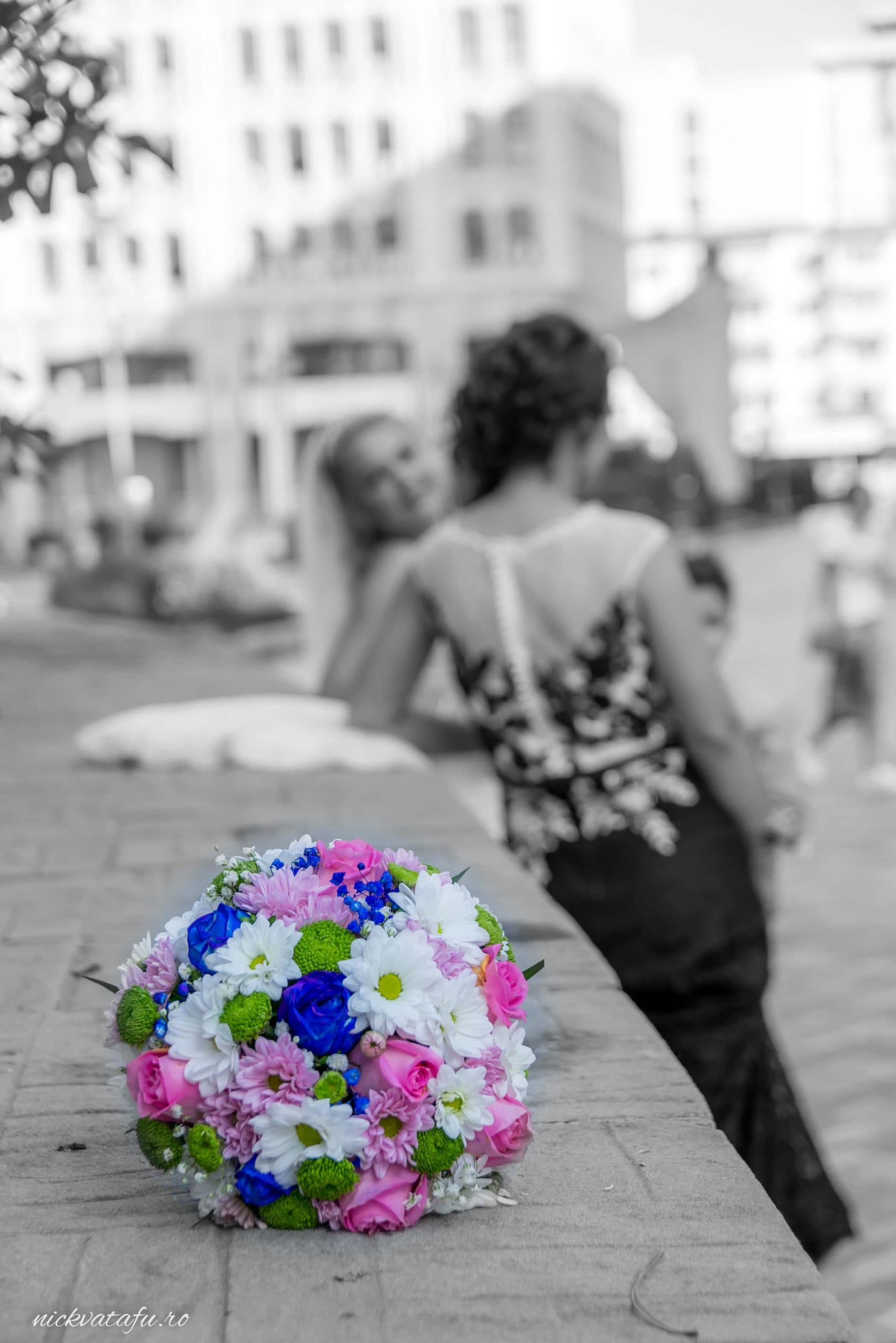 Buchet colorat de flori în prim-plan, cu două femei zâmbind în fundal, fotografie de nuntă plină de emoție.