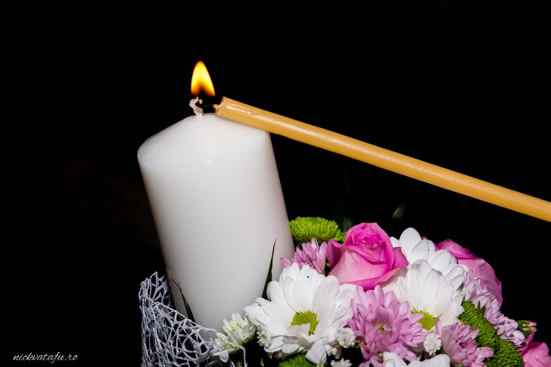 Candelă aprinsă alături de flori delicate, simbolizând emoția unei ceremonii de nuntă sau botez.