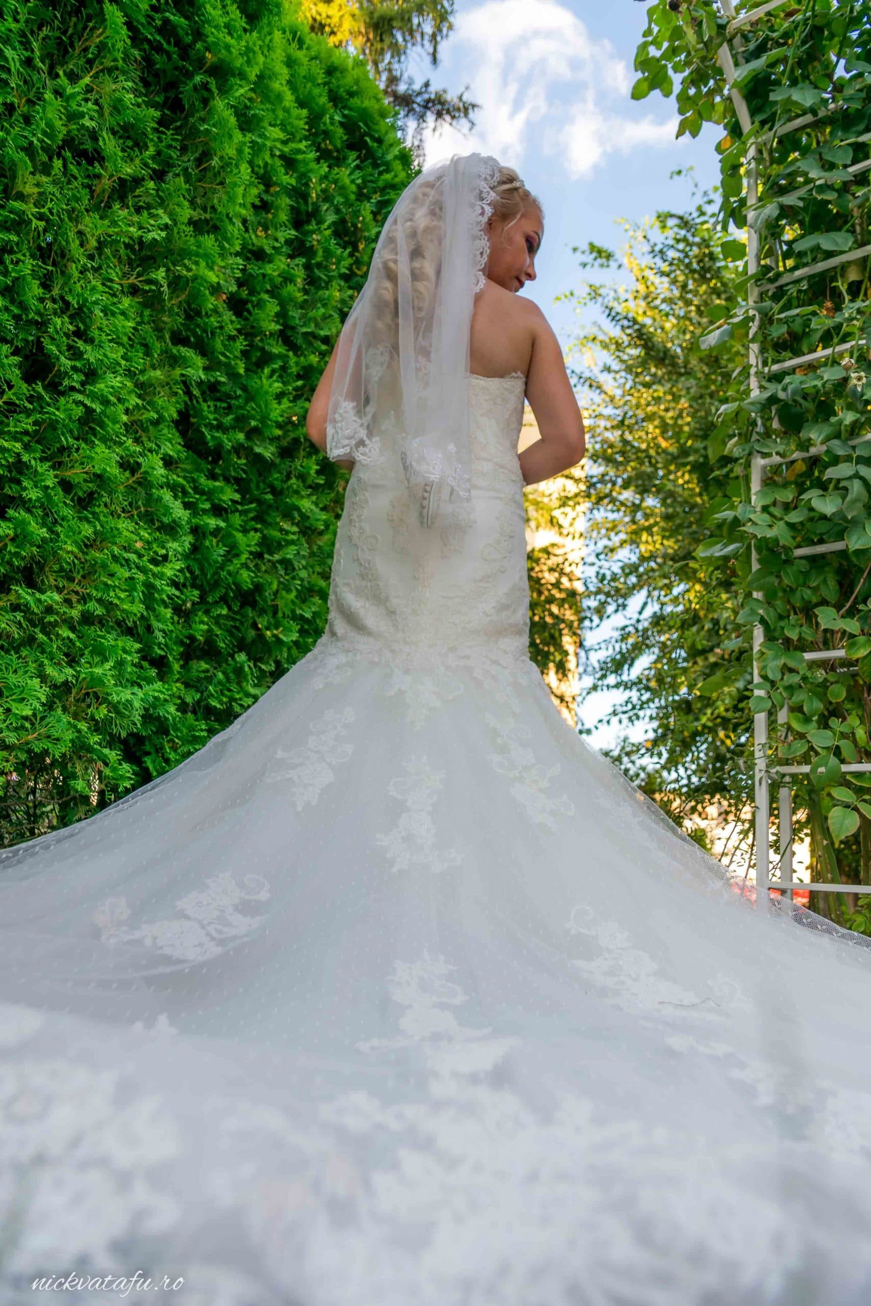 Mireasă elegantă în rochie cu trenă, surprinsă în lumina soarelui, detalii nuntă într-un cadru natural.