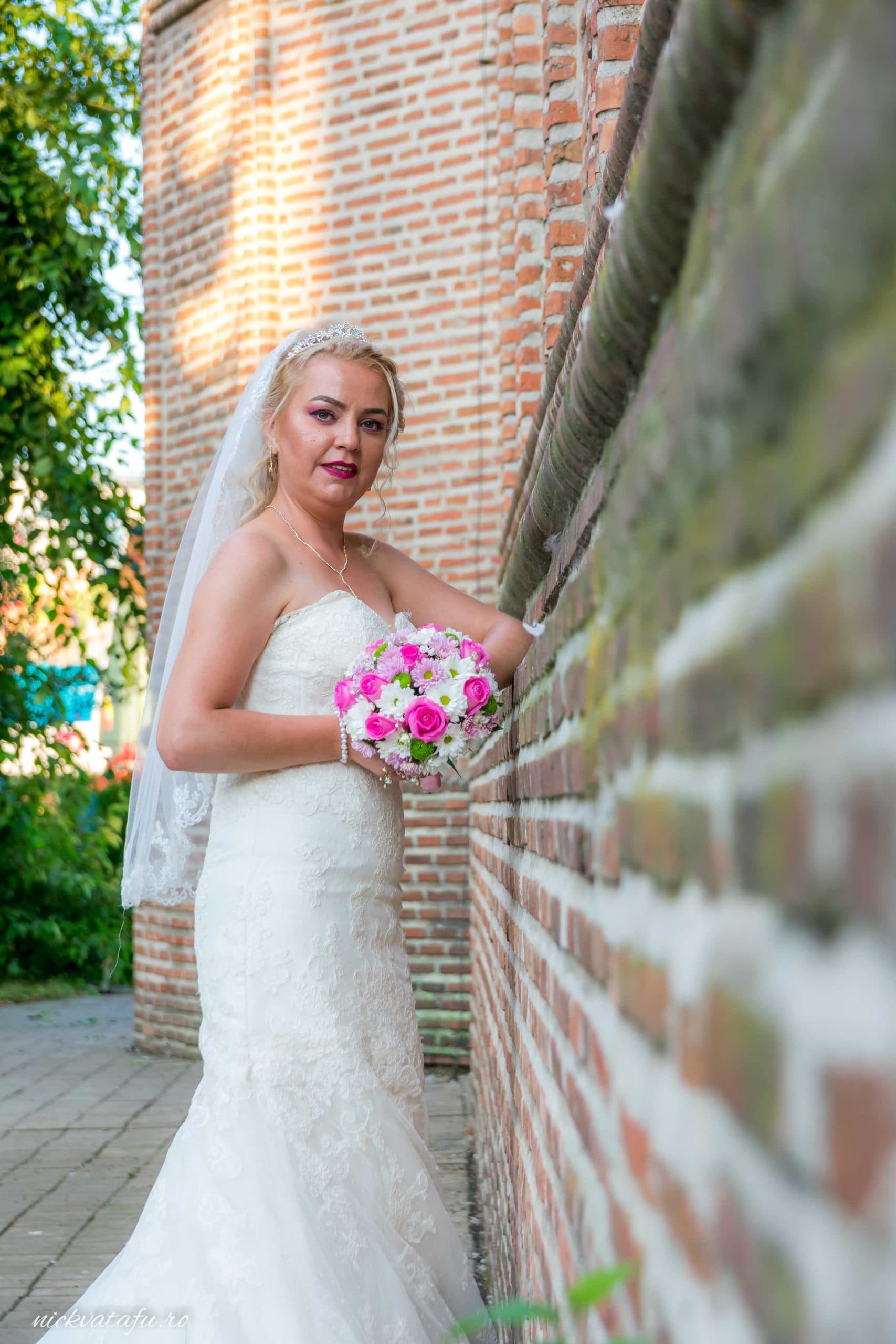 Mireasă zâmbitoare, cu buchet de flori, sprijinită de un zid de cărămidă, capturând emoția unei zile speciale.
