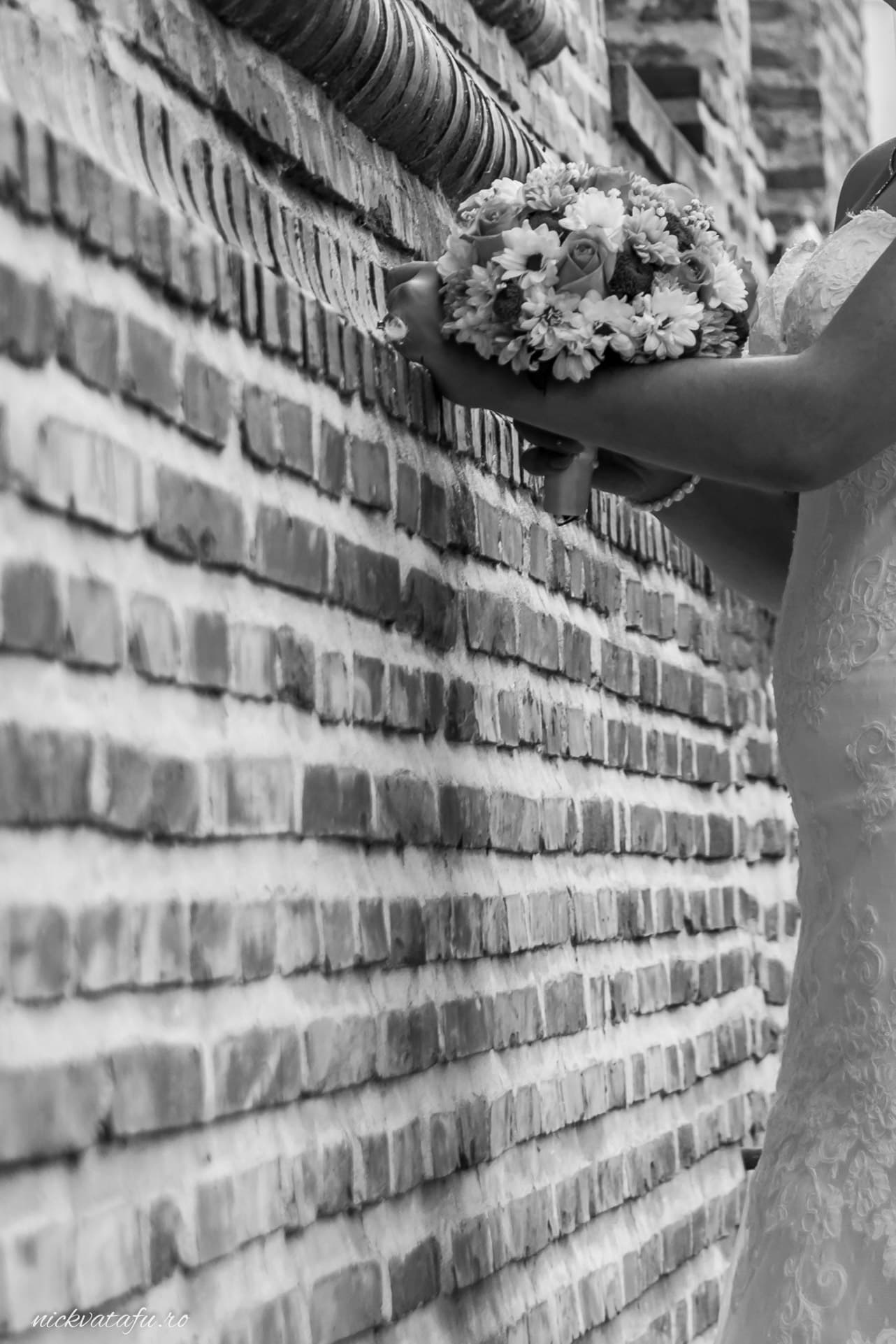 Mireasă elegantă ținând un buchet de flori lângă un zid din cărămidă, fotografie nuntă plină de emoție.