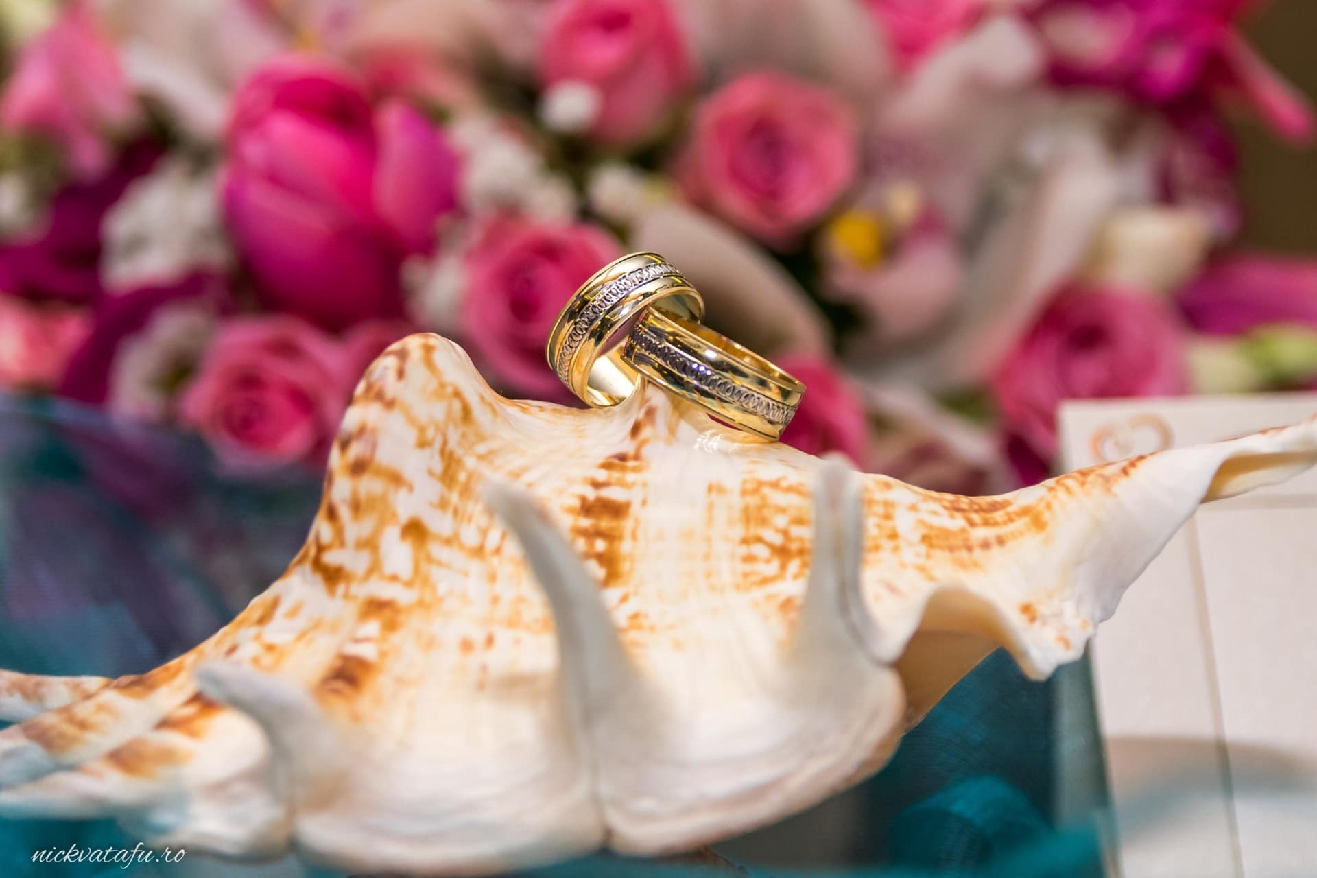 Inel de logodnă așezat pe o scoică, cu flori roz de fundal, detaliu elegant dintr-o fotografie de nuntă.