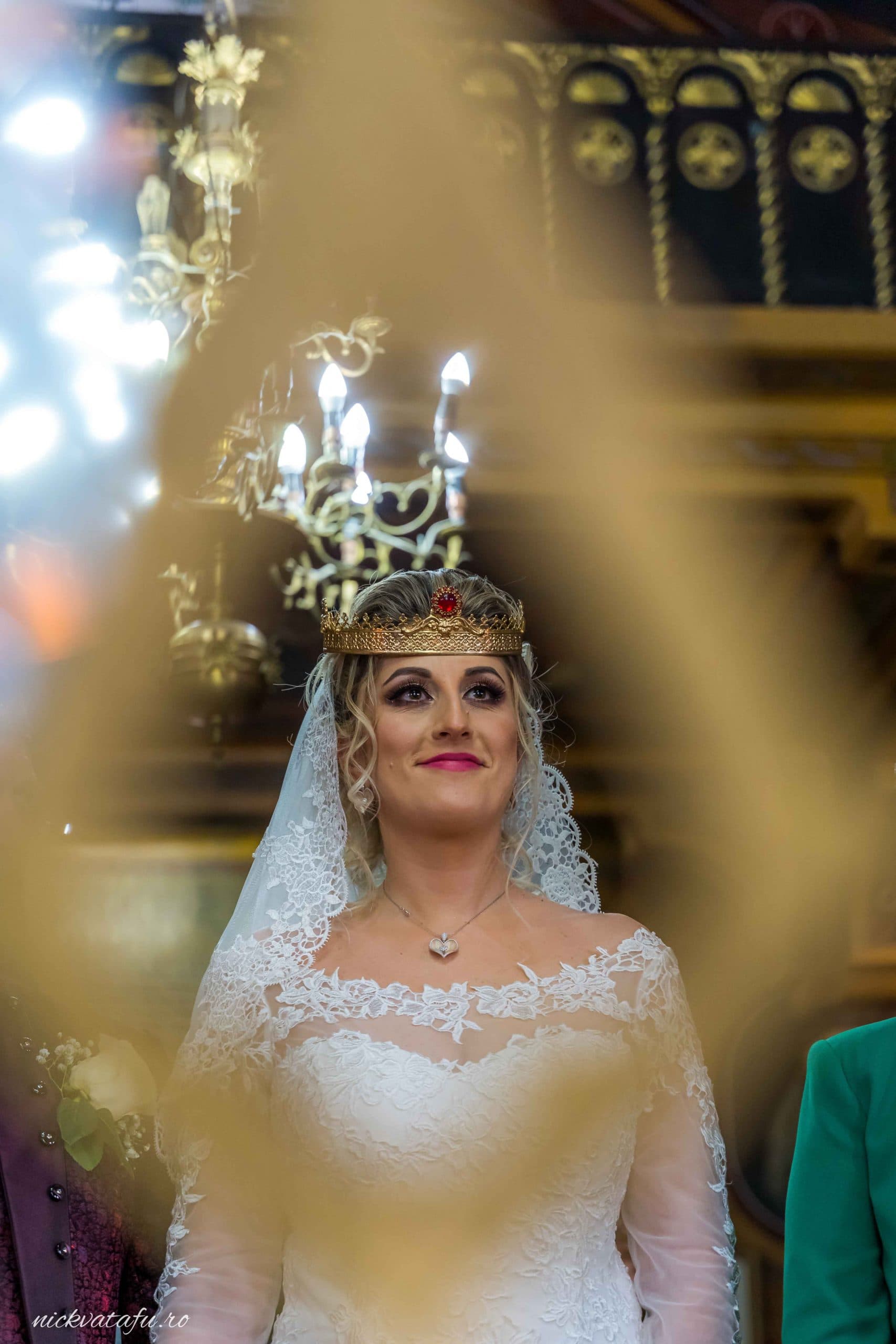 Mireasă cu coroana, zâmbind emoționată în timpul ceremoniei, detalii nuntă în lumina candelabrelor.