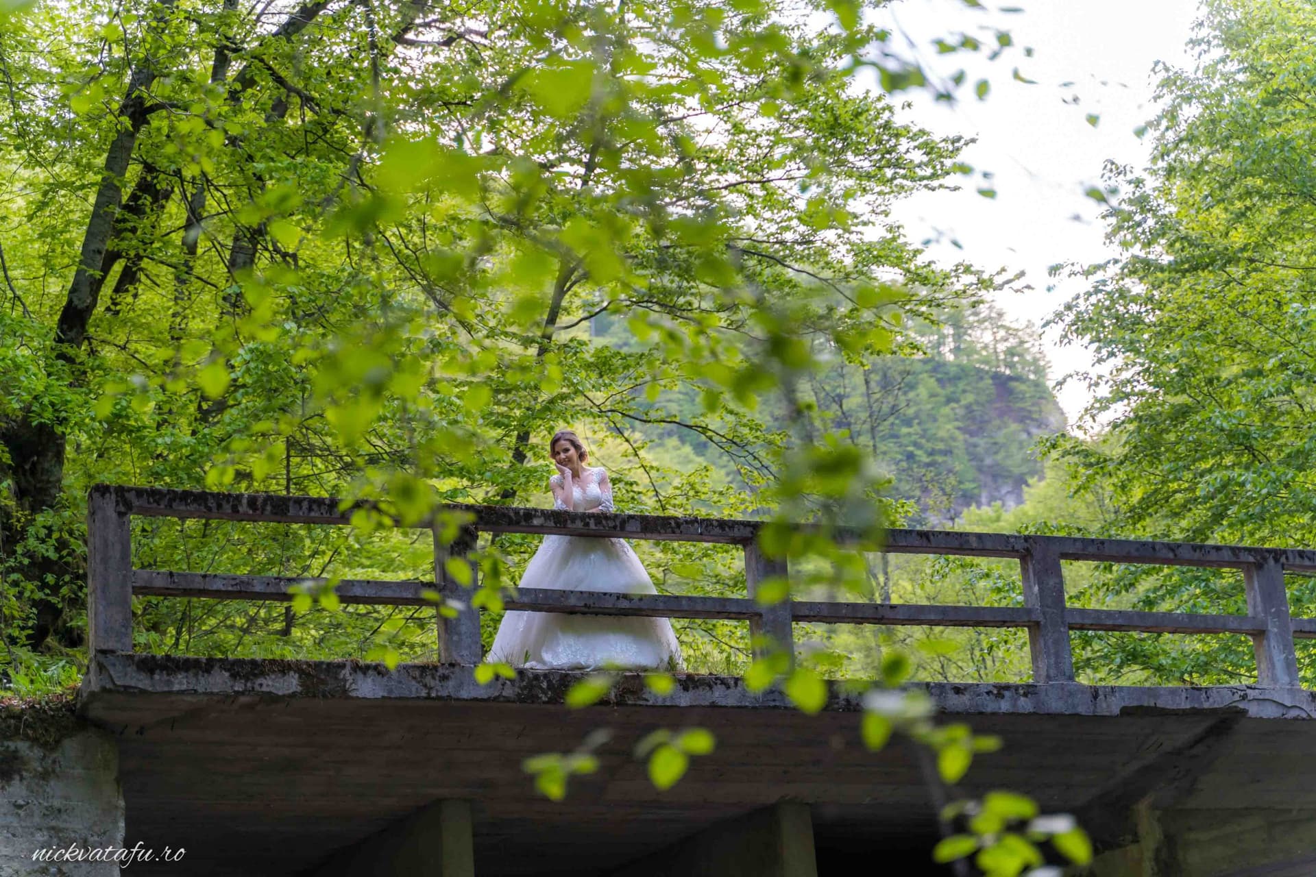 Mireasă contemplativă pe un pod înconjurat de natură, fotografie de nuntă plină de emoție și frumusețe.