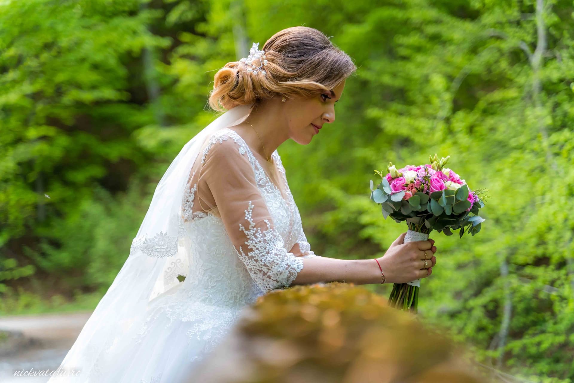 Mireasă contemplativă, cu buchet de flori, într-un peisaj natural plin de verdeață, fotografie de nuntă elegantă.