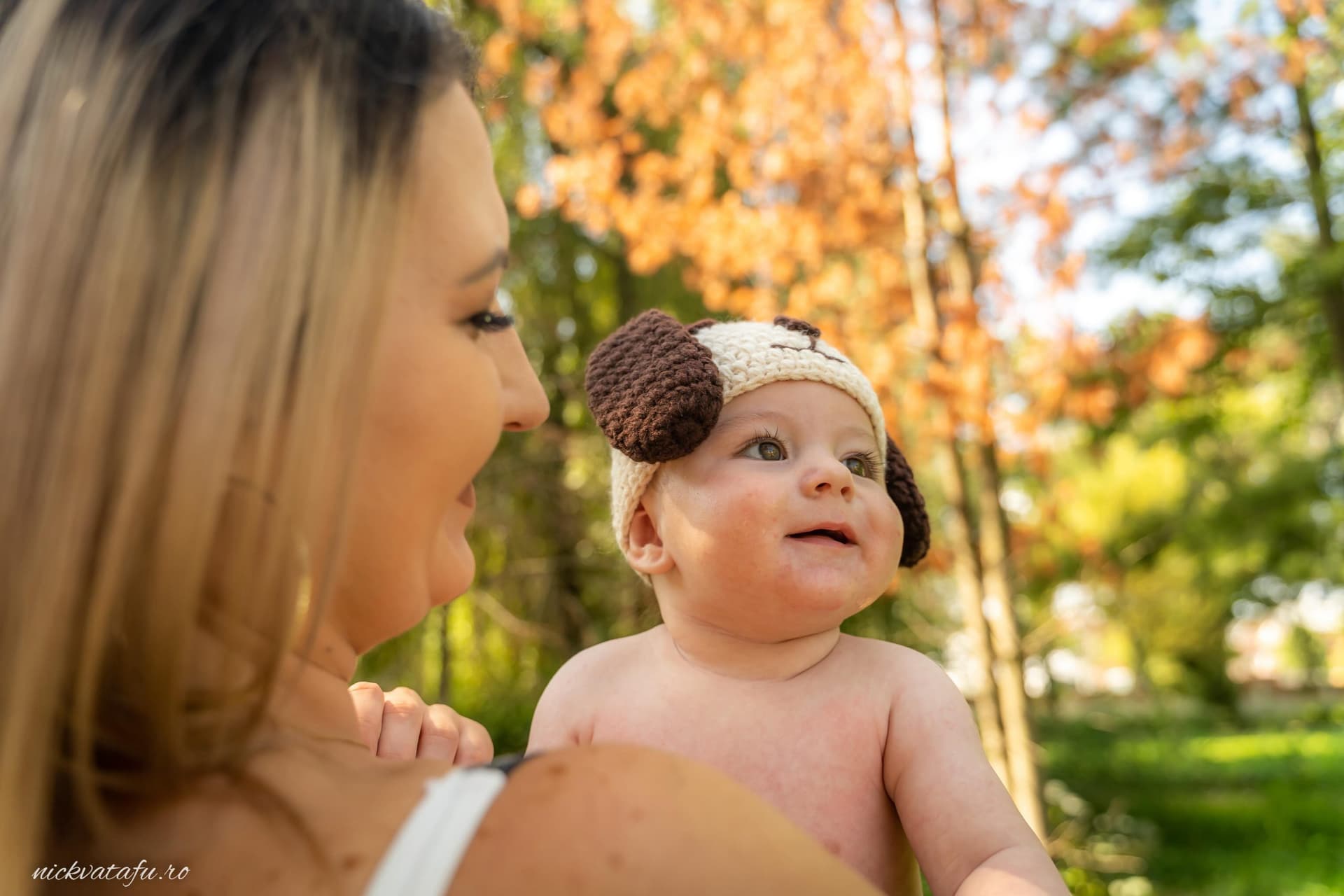Bebeluș zâmbitor, purtând o pălărie adorabilă, în brațele mamei, în mijlocul naturii, evocând emoție și bucurie.