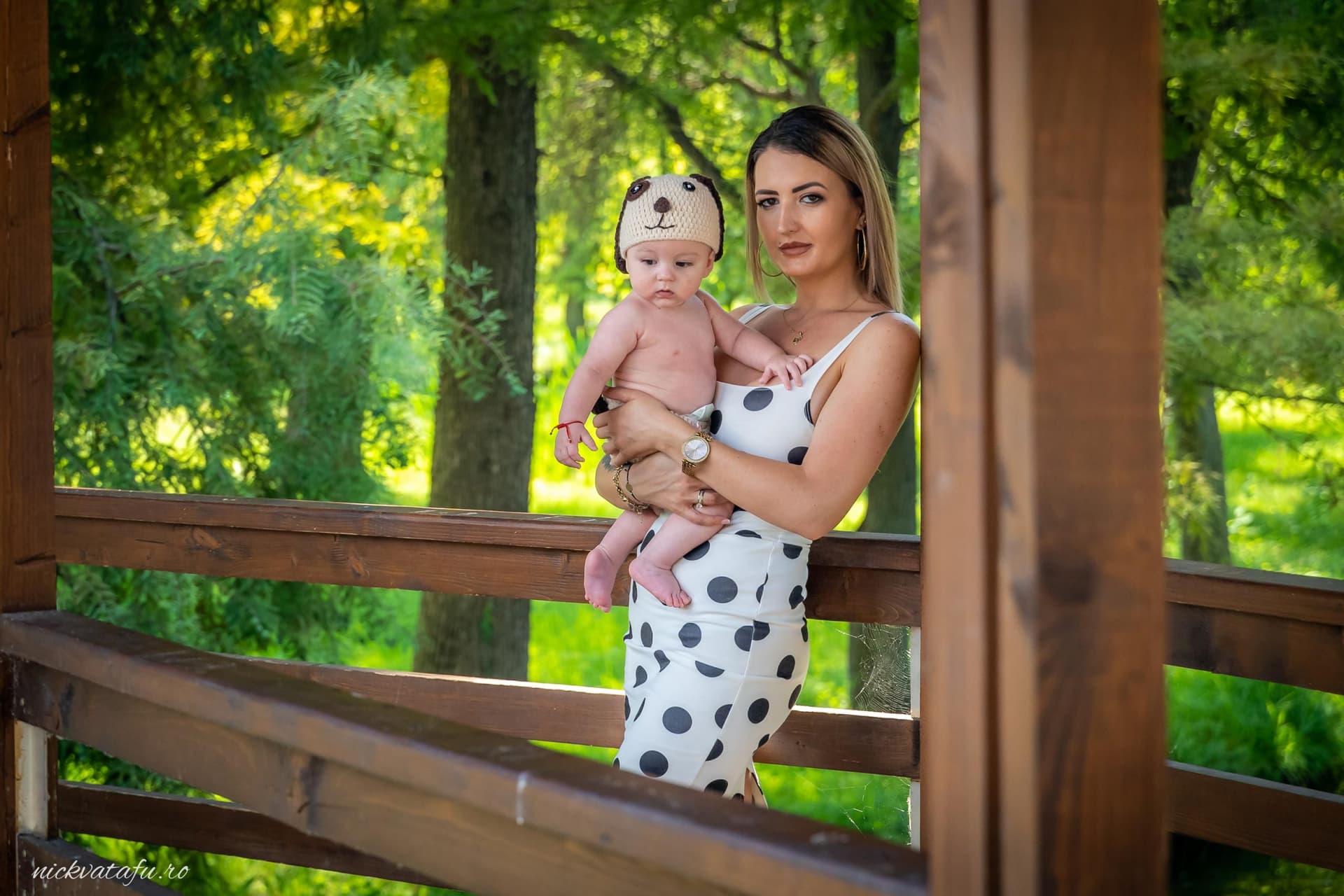 Mamă îmbrăcată elegant, ținând un bebeluș pe un balcon, înconjurată de natură verde, fotografie emoționantă de familie.