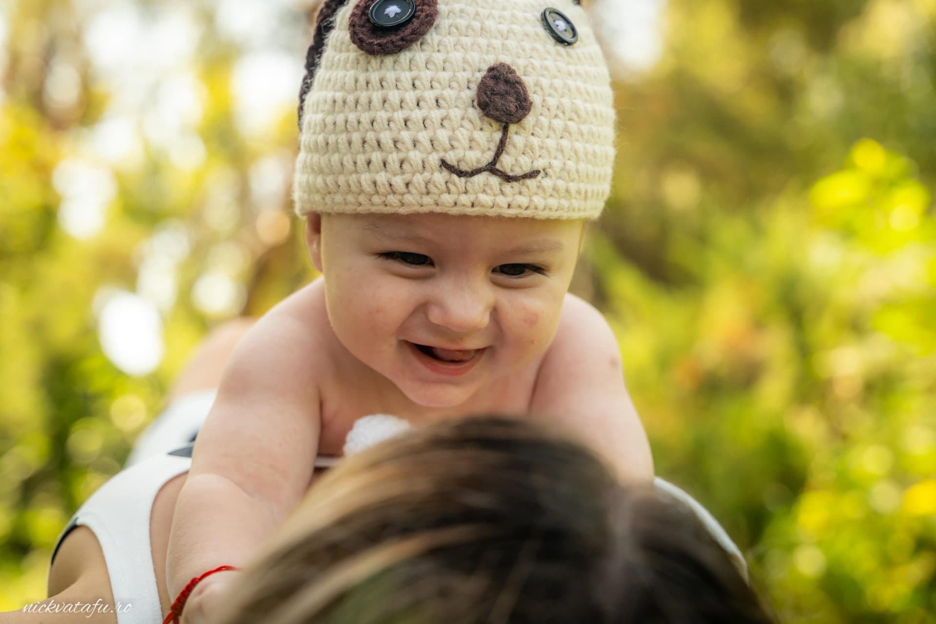 Bebeluș zâmbitor cu pălărie amuzantă, jucându-se cu mama într-un cadru natural plin de emoție.