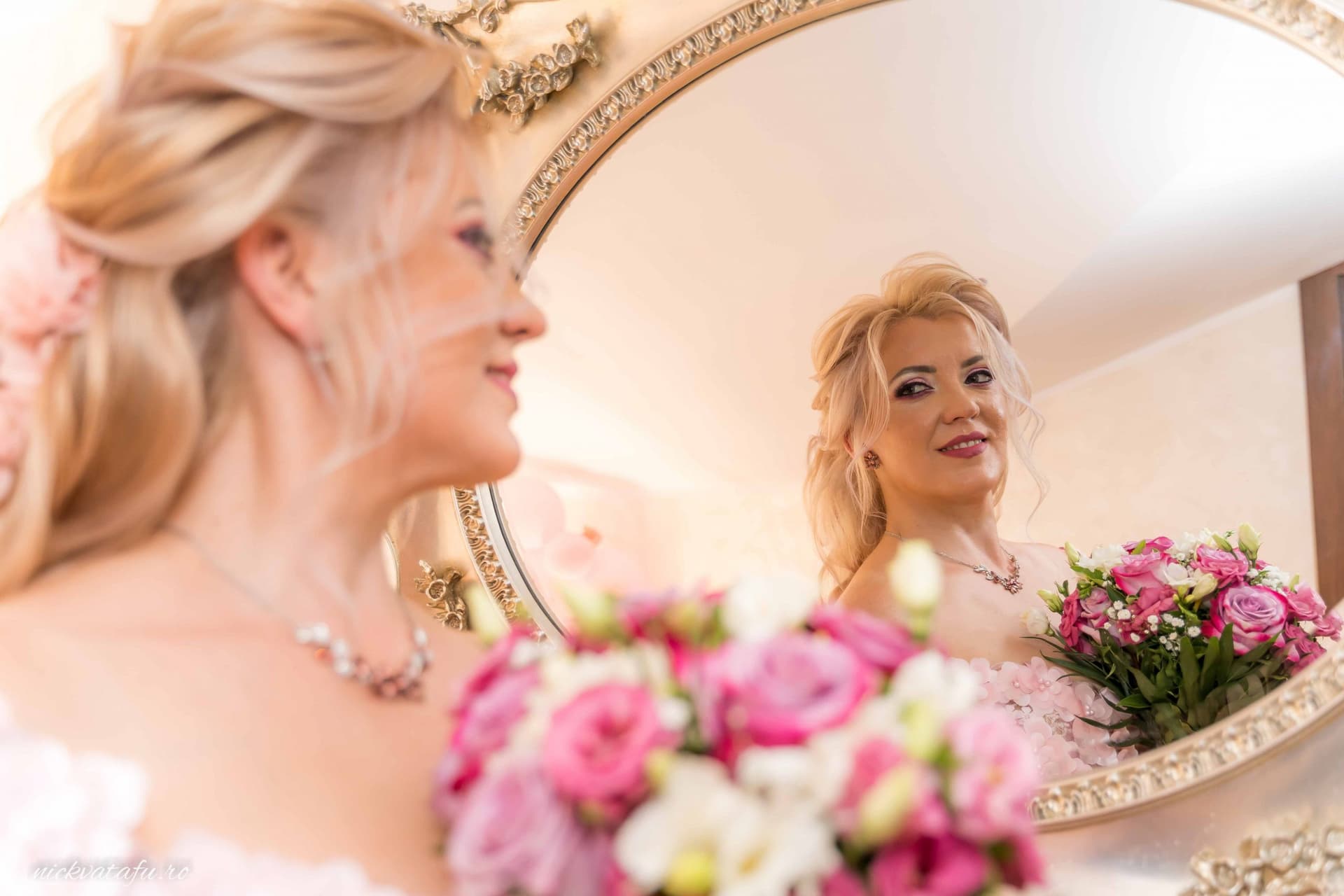 Mireasă zâmbitoare, admirându-se în oglindă cu un buchet de flori, detalii nuntă elegante și emoție autentică.