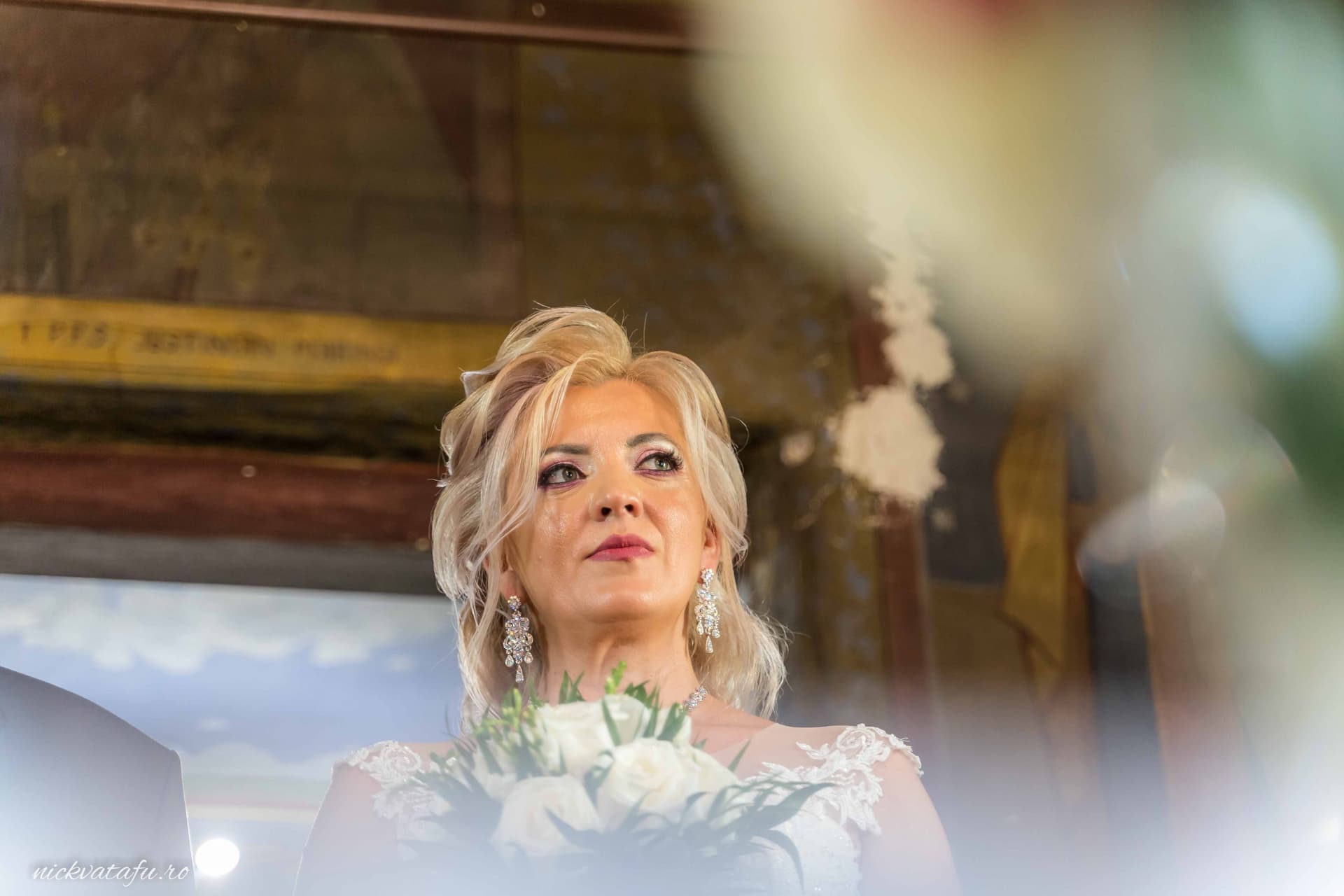 Mireasă elegantă, cu buchet de flori, privind emoționată, într-un cadru de nuntă plin de detalii.