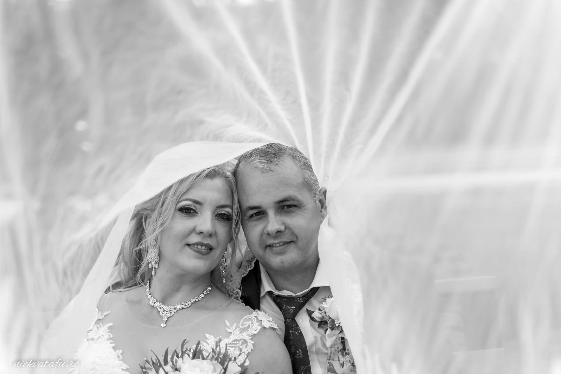 Mire și mireasă zâmbind fericiți, sub voal, emanând emoție și iubire într-o fotografie de nuntă elegantă.