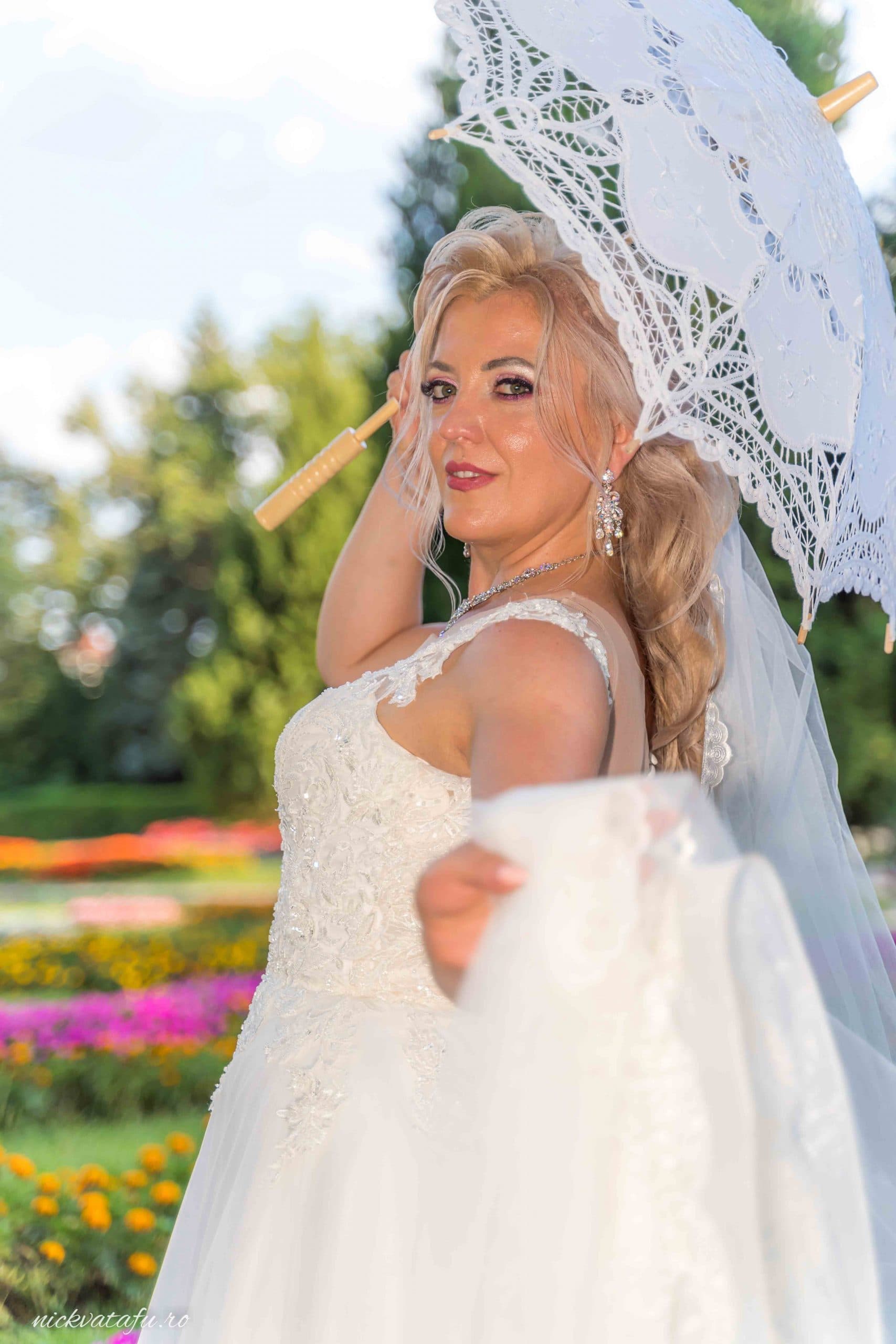 Mireasă elegantă cu umbrela, zâmbind într-o grădină plină de flori, fotografie de nuntă plină de emoție.