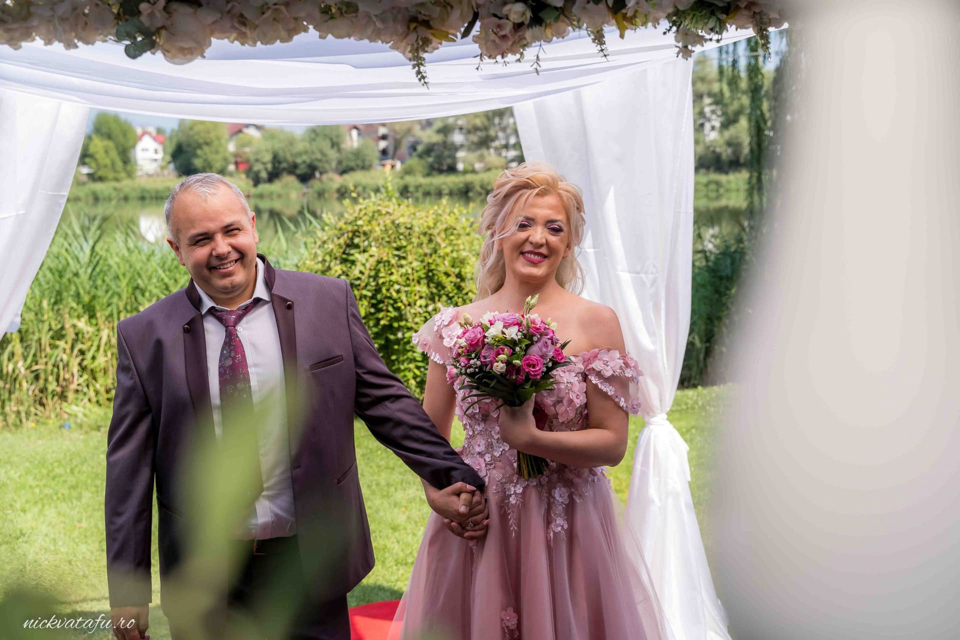 Mire și mireasă zâmbind emoționat, în fața unui decor elegant, fotografie de nuntă realizată lângă apă.