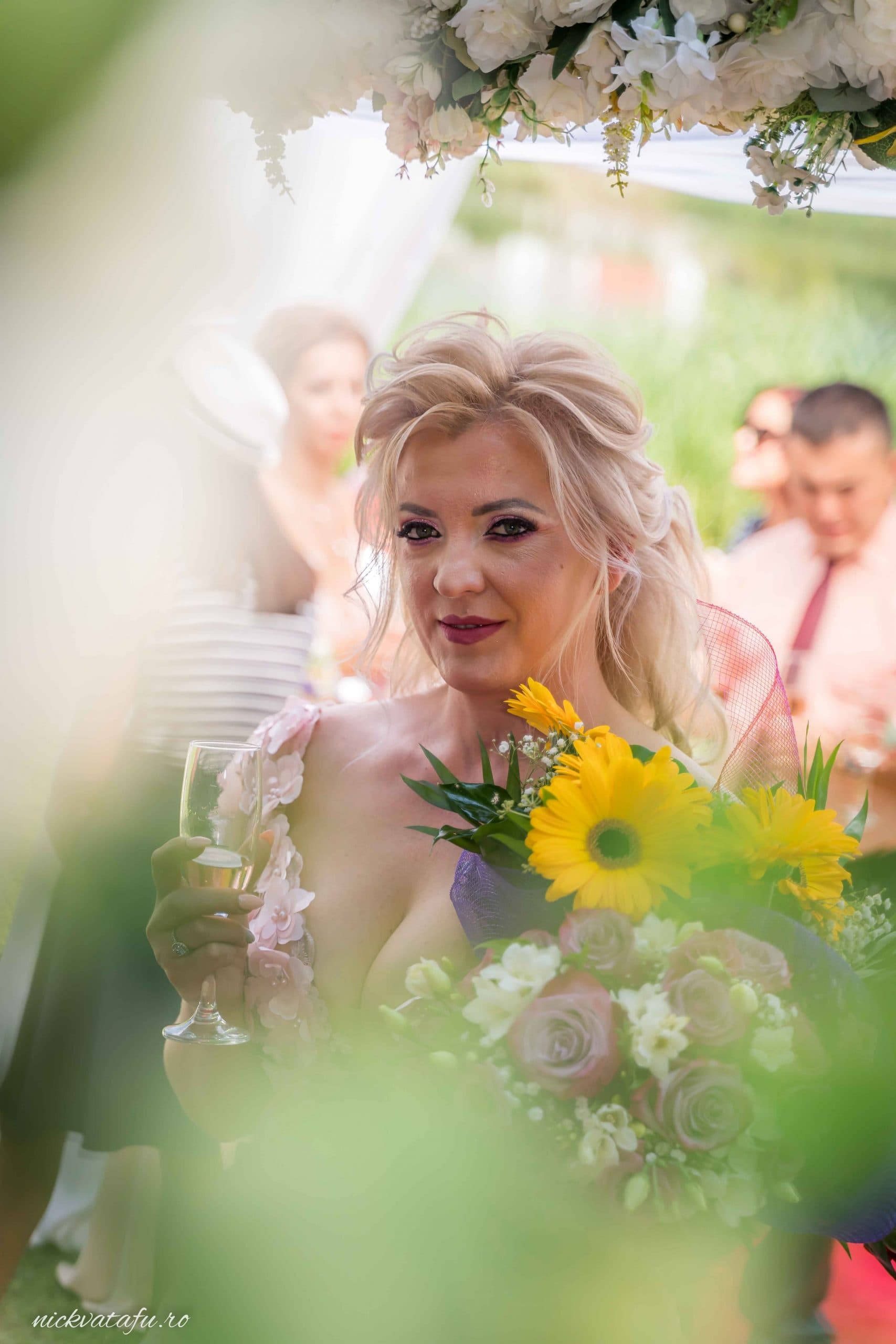 Mireasă elegantă, zâmbind cu buchet de flori, în fundal petrecere veselă, fotografie nuntă plină de emoție.