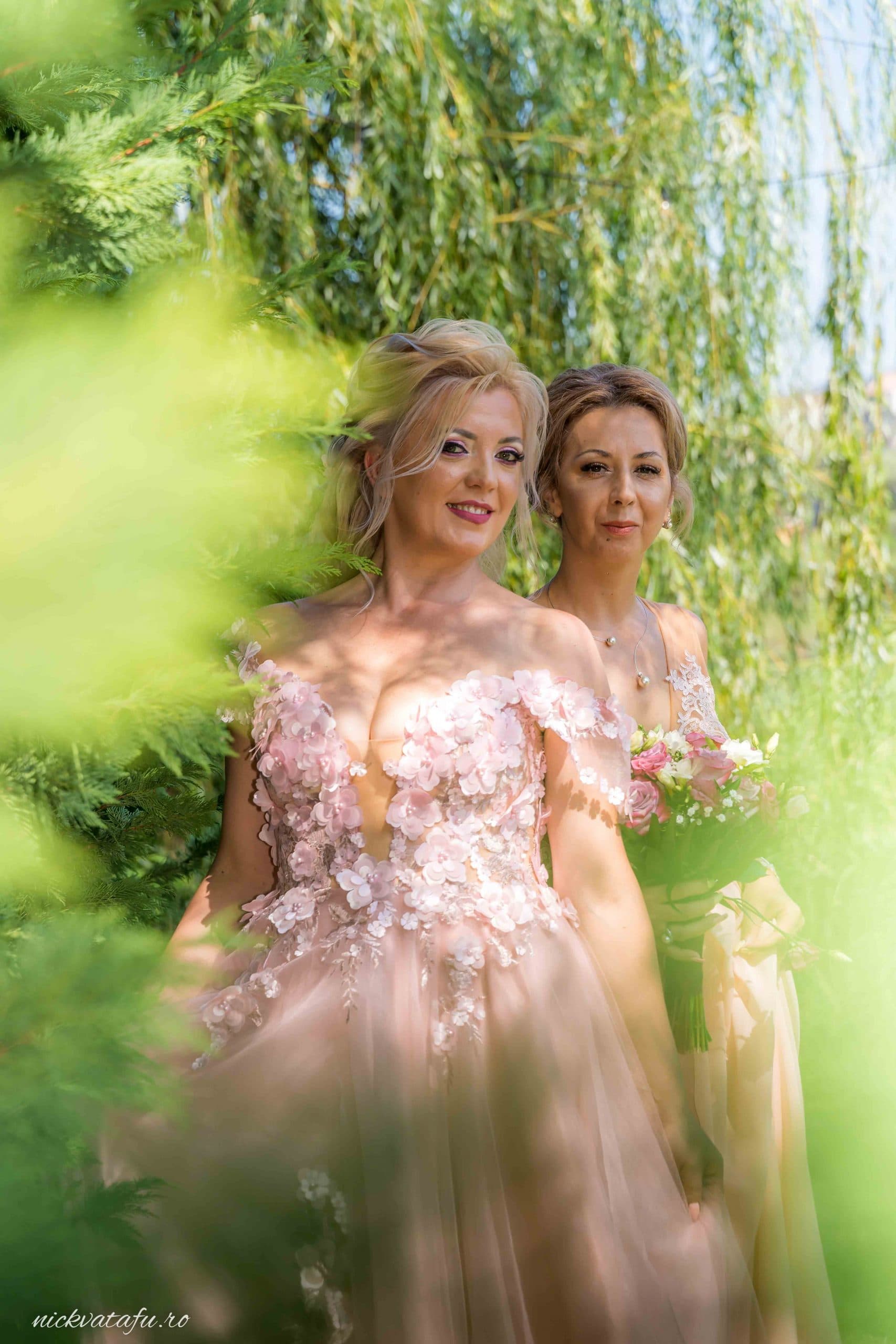 Mireasă și domnișoară de onoare zâmbind printre frunze verzi, fotografie de nuntă plină de emoție și eleganță.