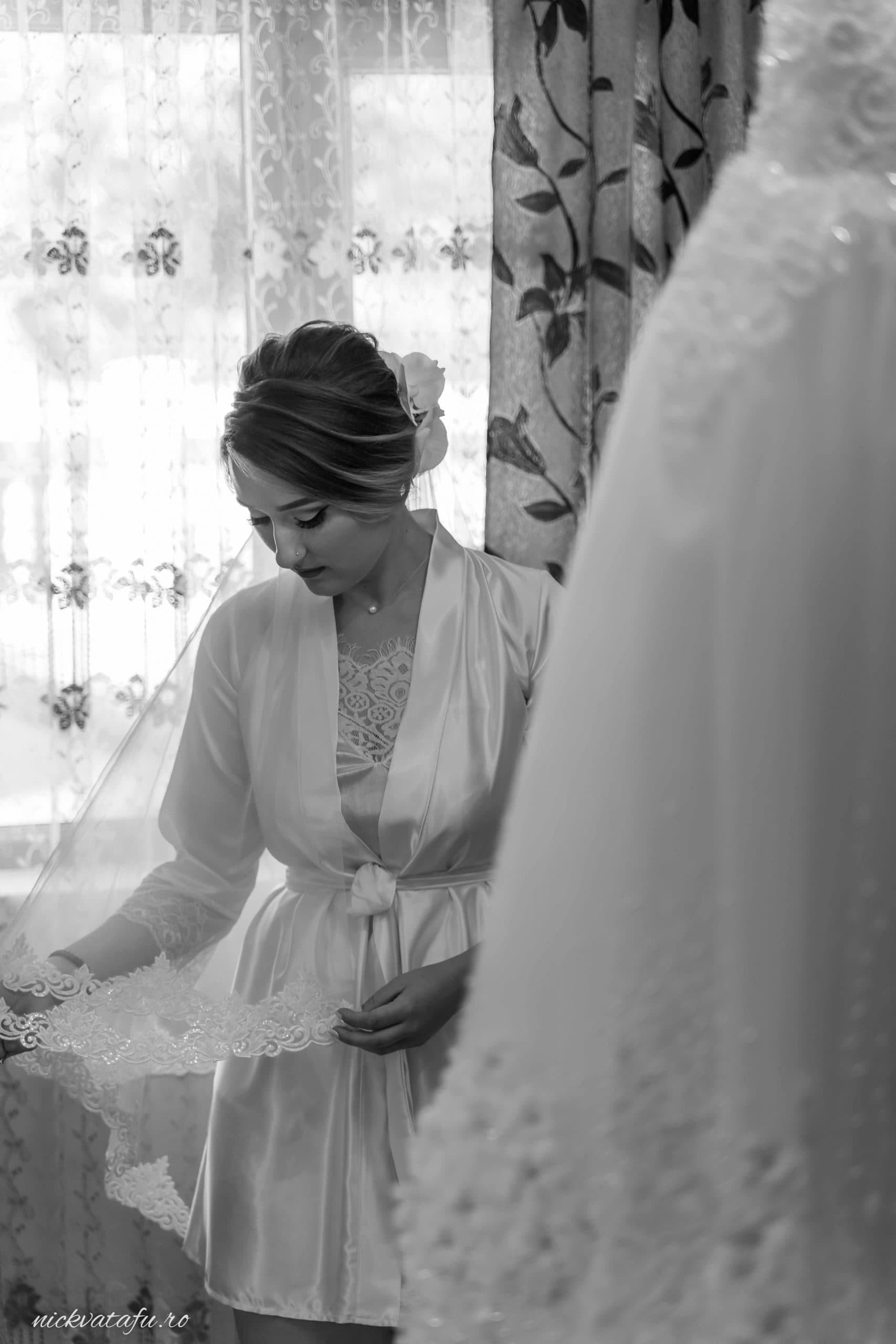 Mireasă contemplând cu emoție rochia de nuntă, lumină naturală filtrată prin perdele, fotografie elegantă de nuntă.