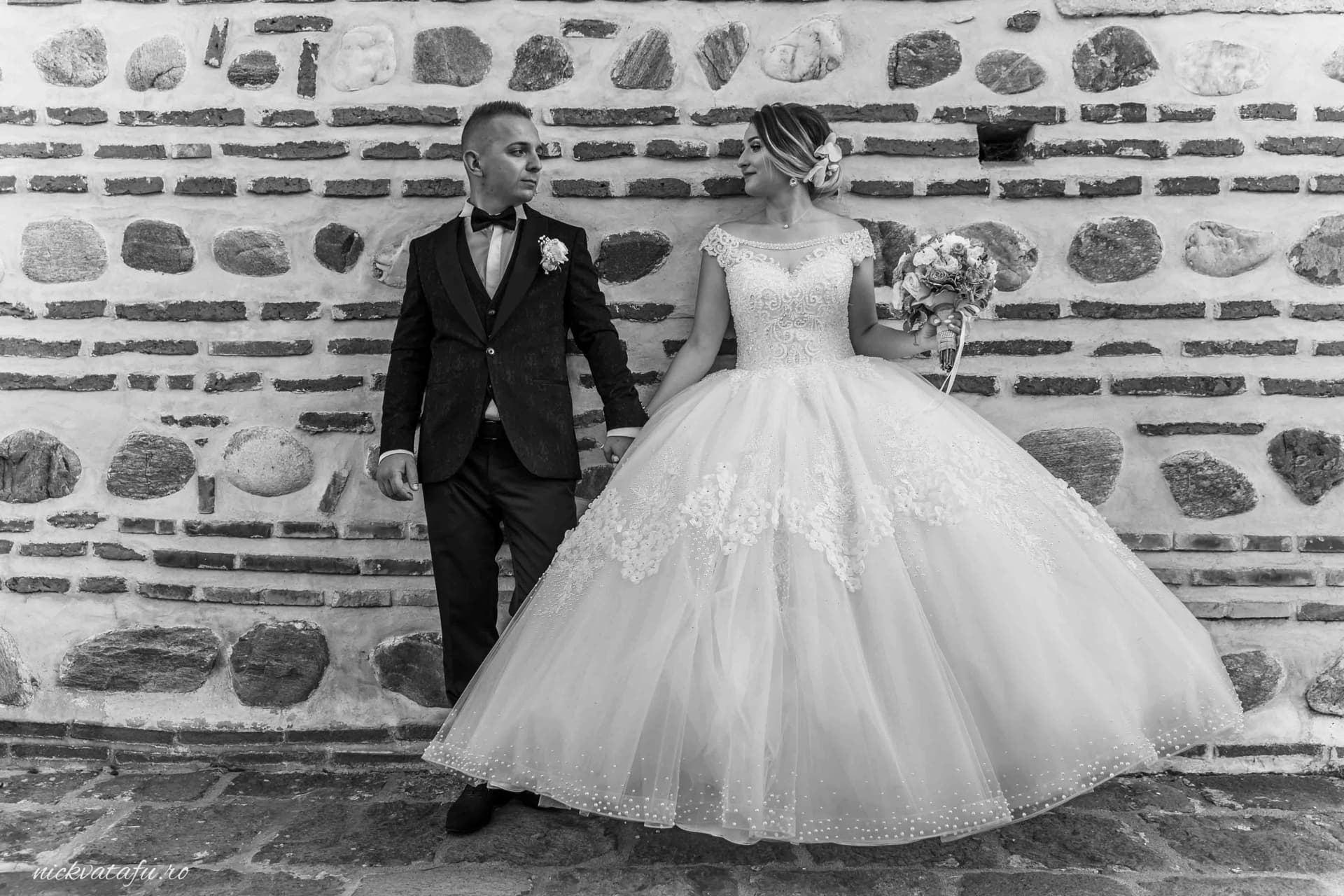 Mire și mireasă, ținându-se de mână, în fața unei pereți din piatră, emanând emoție și eleganță.