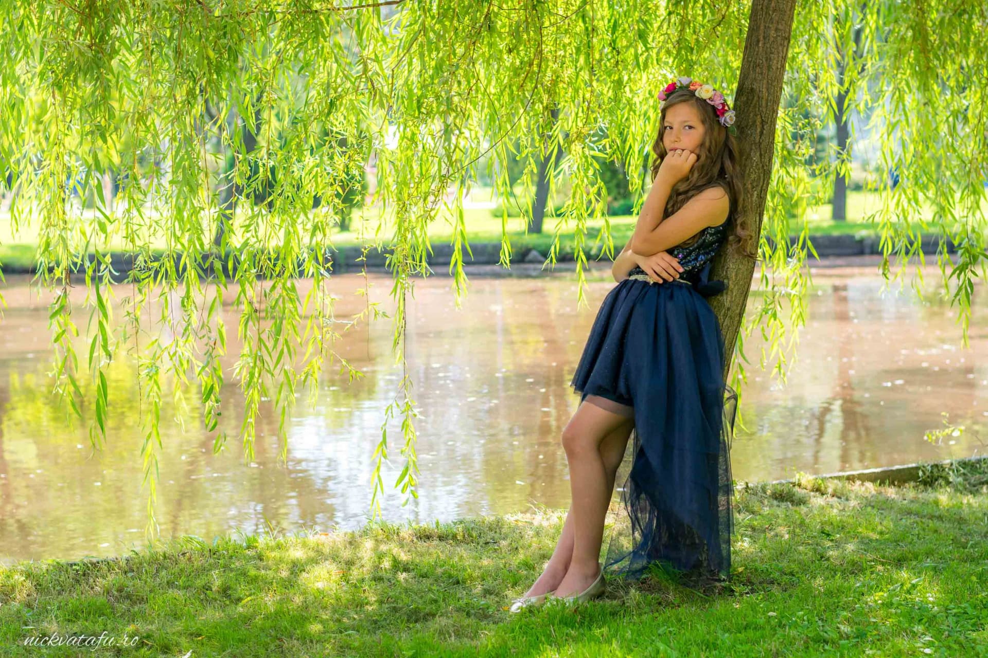 Fată elegantă, cu rochie albastră, stând lângă un copac, pe malul unui lac, emanând emoție și naturalețe.
