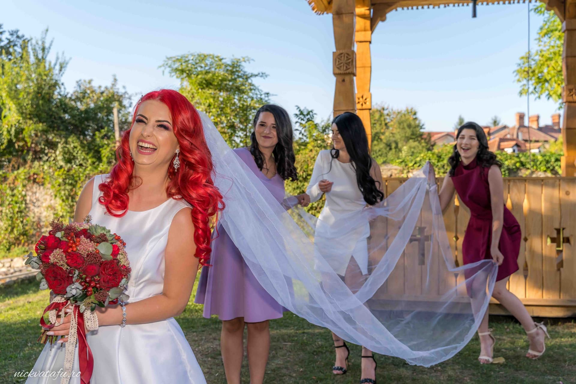 Mireasă cu părul roșu și buchet de flori, însoțită de domnițe de onoare, zâmbind la o nuntă în aer liber.