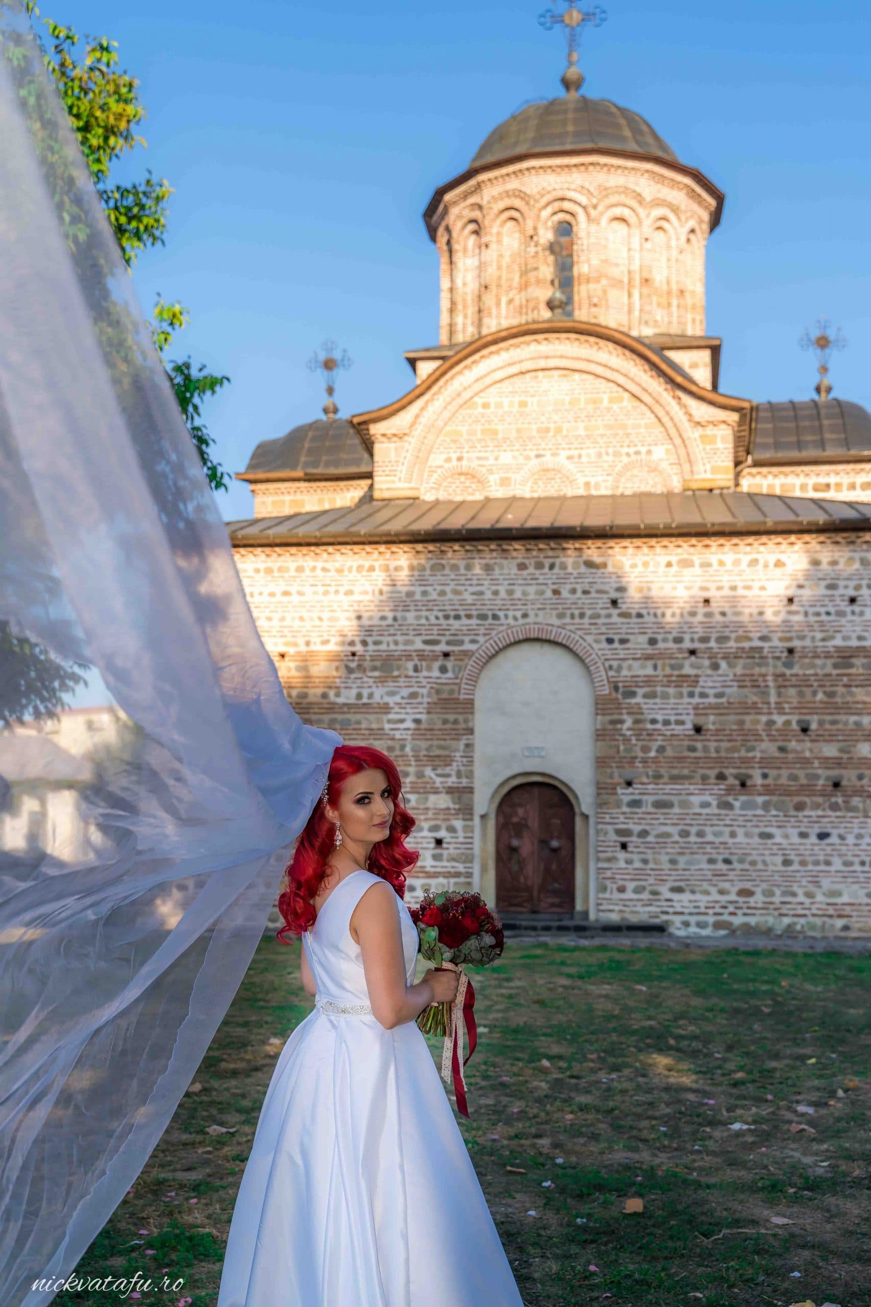 Mireasă cu părul roșu, zâmbind și ținând buchetul, în fața unei biserici istorice, fotografie de nuntă plină de emoție.