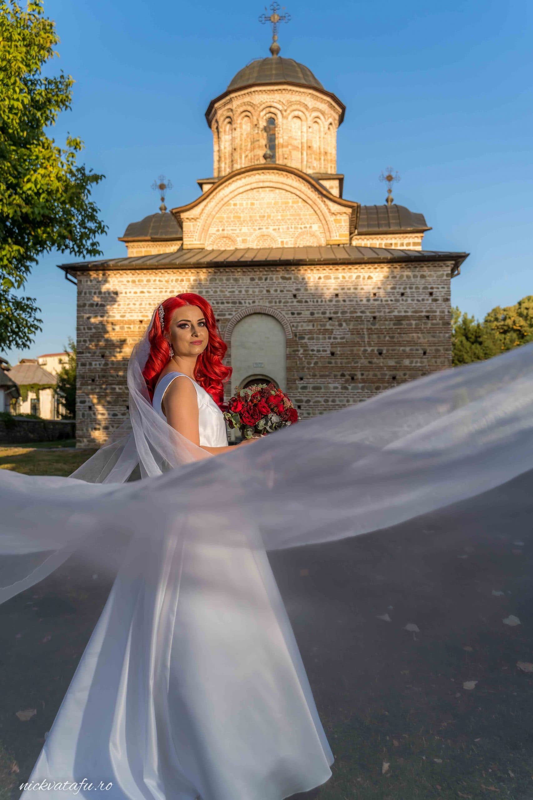 Mireasă cu voal și buchet, zâmbind fericită în fața unei biserici, fotografie de nuntă plină de emoție.