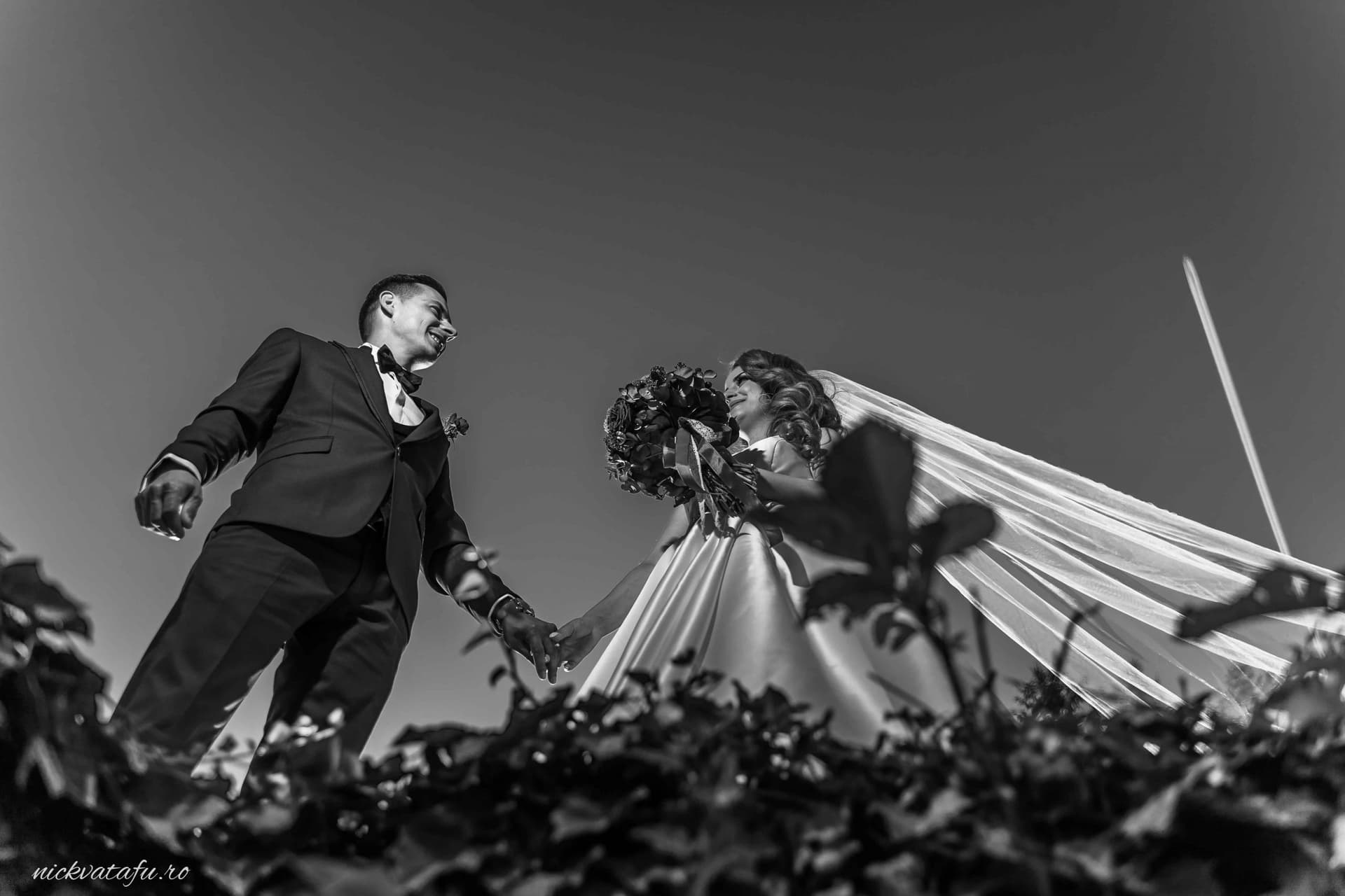 Mire și mireasă zâmbind și ținându-se de mână, fotografie nuntă în alb-negru cu detalii elegante în fundal.