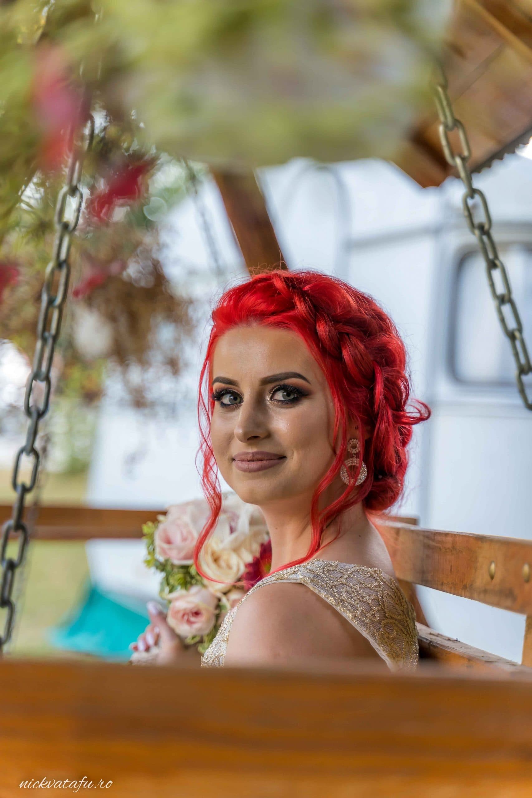 Mireasă cu părul roșu, zâmbind pe o balansoar, cu flori în brațe, fotografie de nuntă plină de emoție.
