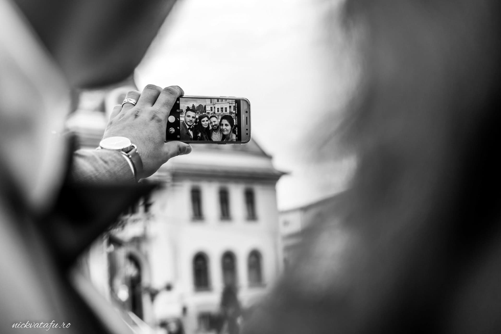 Mână care ține un telefon pentru a captura un moment special, fotografie de eveniment în fundal urban.