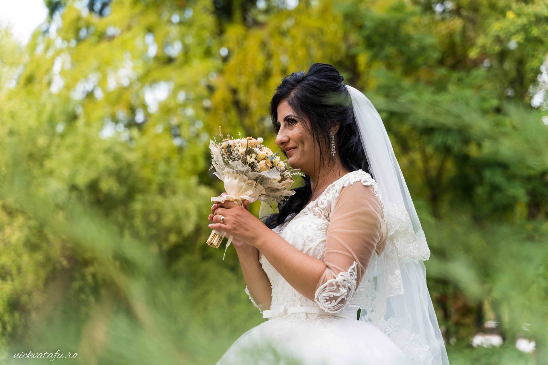 Mireasă zâmbitoare în natură, ținând un buchet de flori, fotografie de nuntă plină de emoție și eleganță.