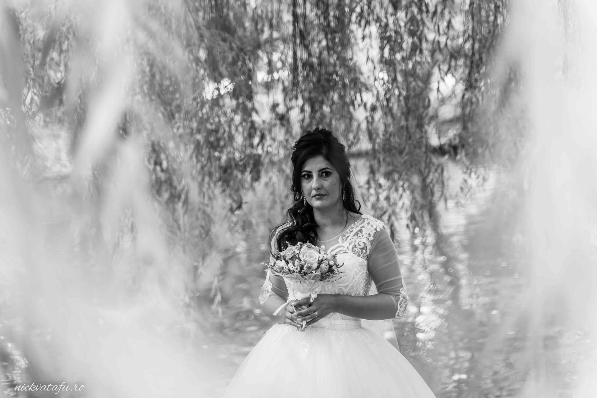 Mireasă elegantă, cu buchet în mână, în fața unui peisaj de apă, fotografie nuntă plină de emoție.