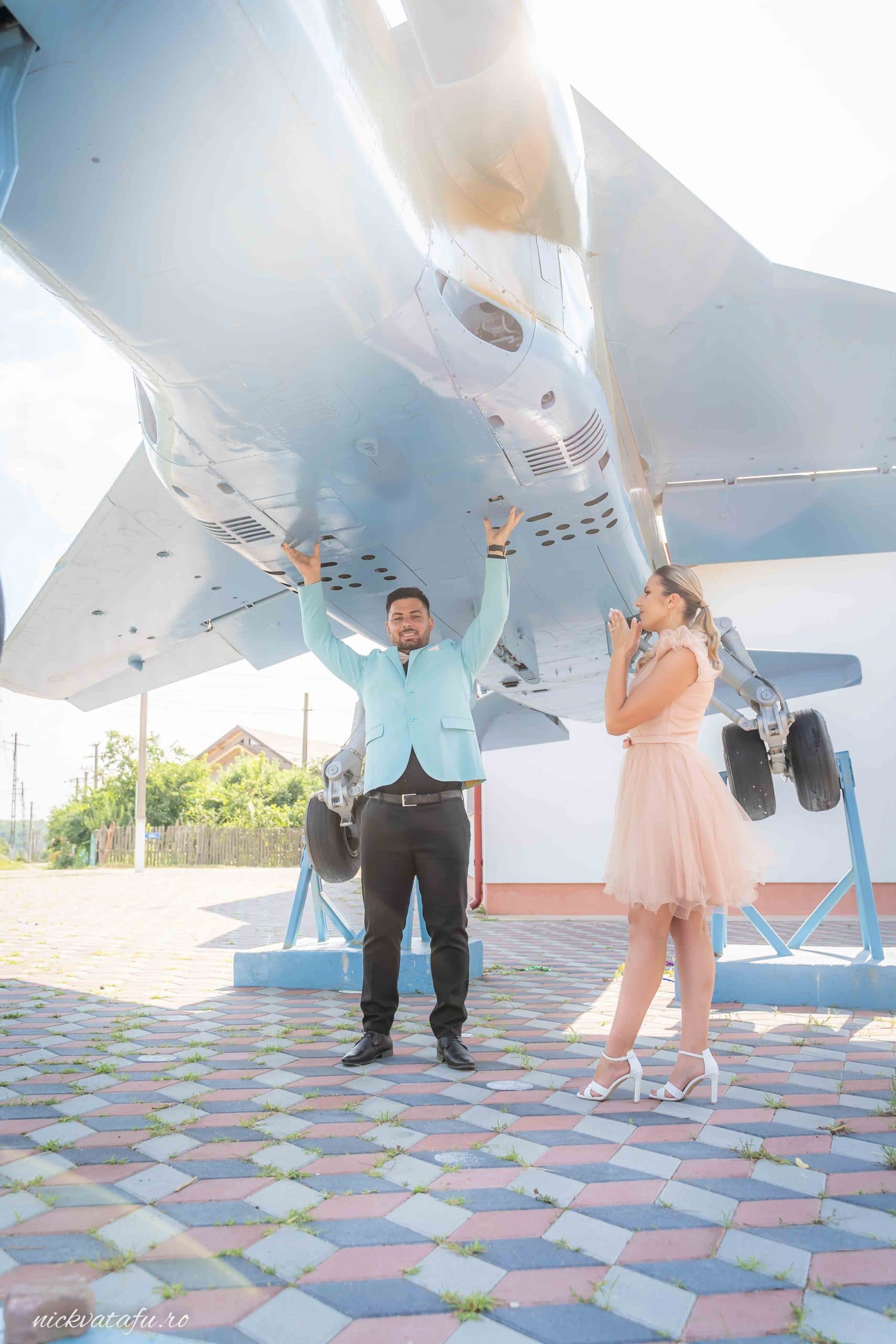 Cuplu fericit pozând sub un avion, într-un cadru neobișnuit, fotografie de nuntă plină de emoție și creativitate.