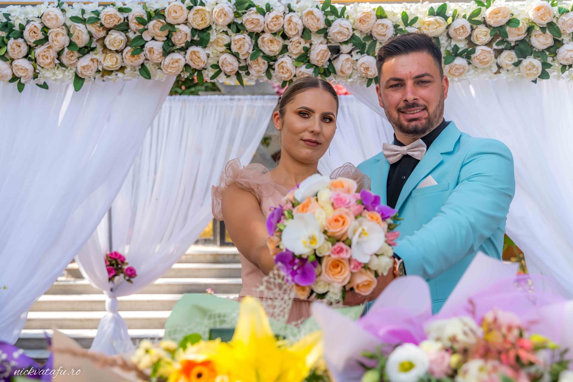 Mire și mireasă zâmbind fericiți, cu buchet colorat, în decor floral elegant, fotografie de nuntă plină de emoție.