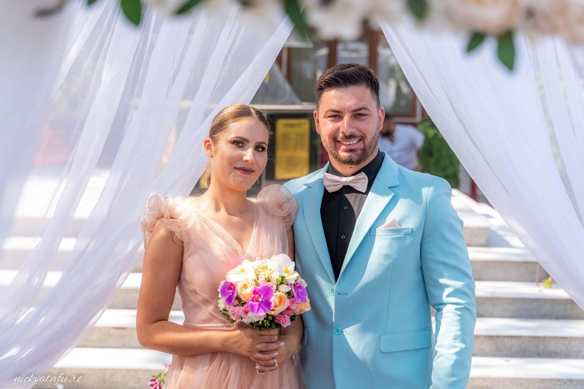 Mire și mireasă zâmbind fericiți, înconjurați de flori, la o ceremonie elegantă de nuntă.