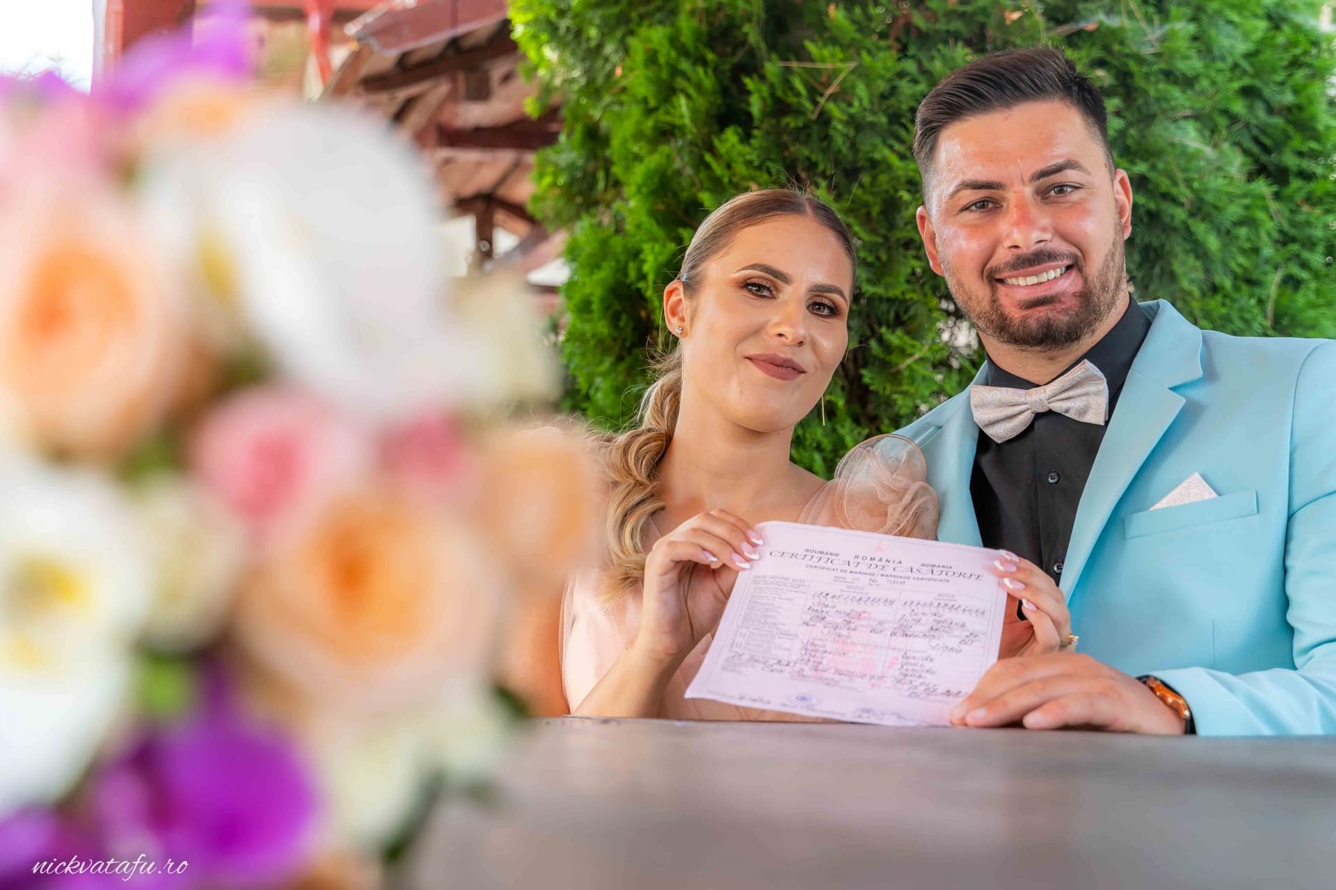 Mire și mireasă zâmbind fericiți, ținând un certificat de căsătorie, în decor floral elegant.