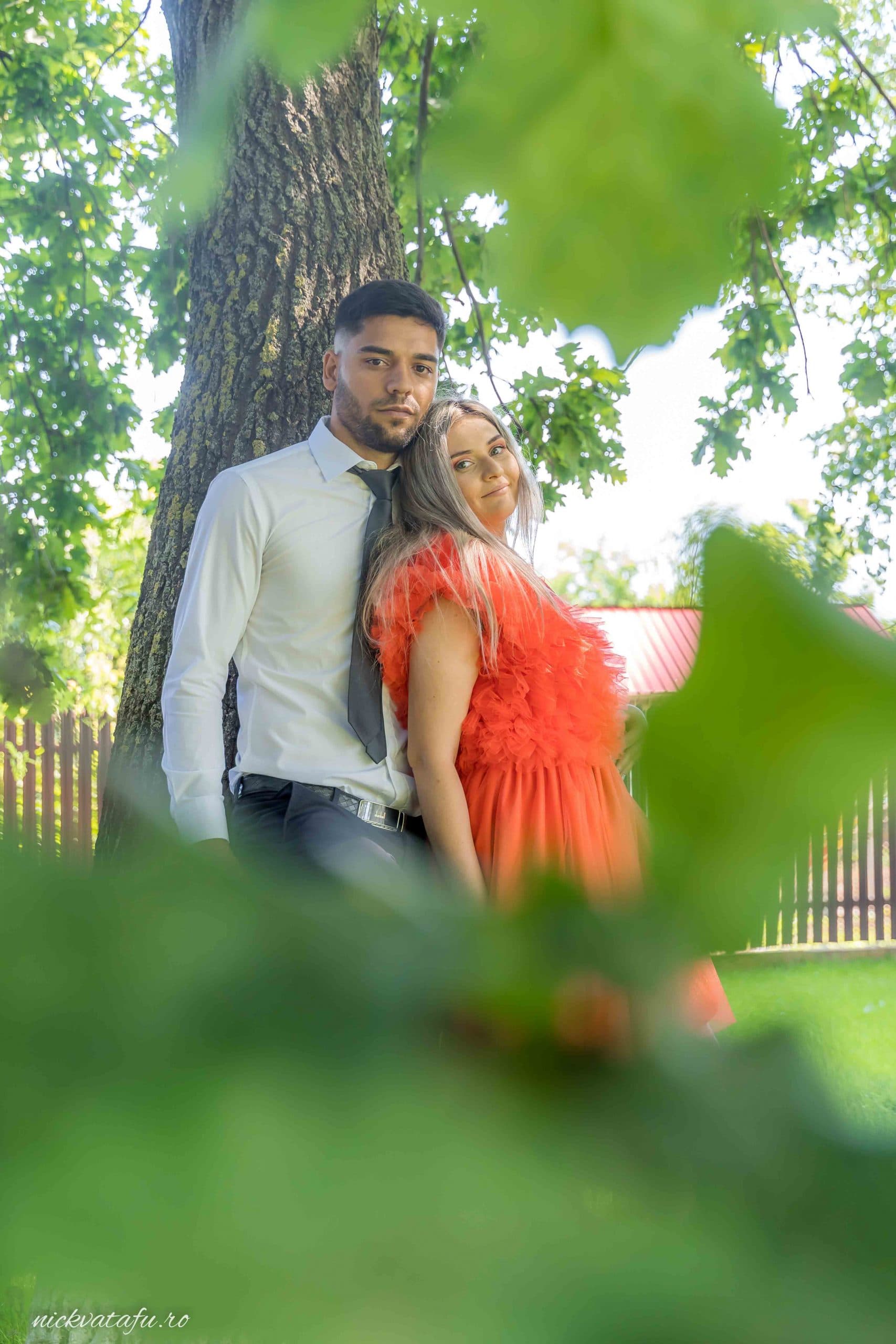 Cuplu îmbrăcat elegant, zâmbind și îmbrățișându-se sub un copac, fotografie nuntă plină de emoție.