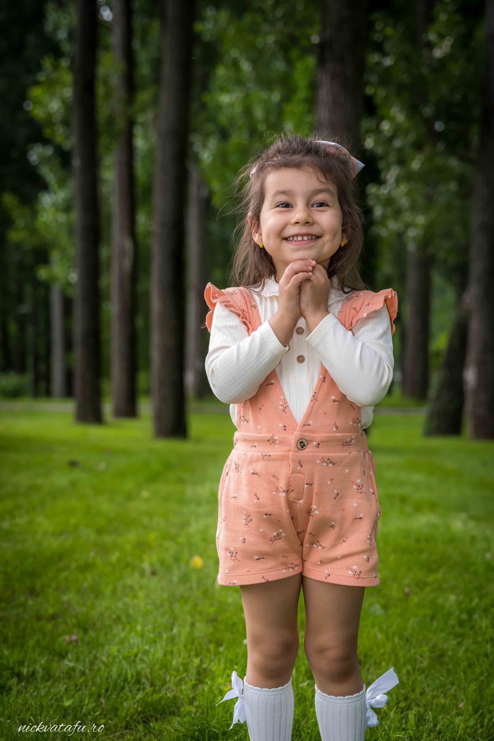 Fetiță zâmbitoare, îmbrăcată în rochiță, stând pe iarbă verde, surprinsă într-un moment de bucurie.