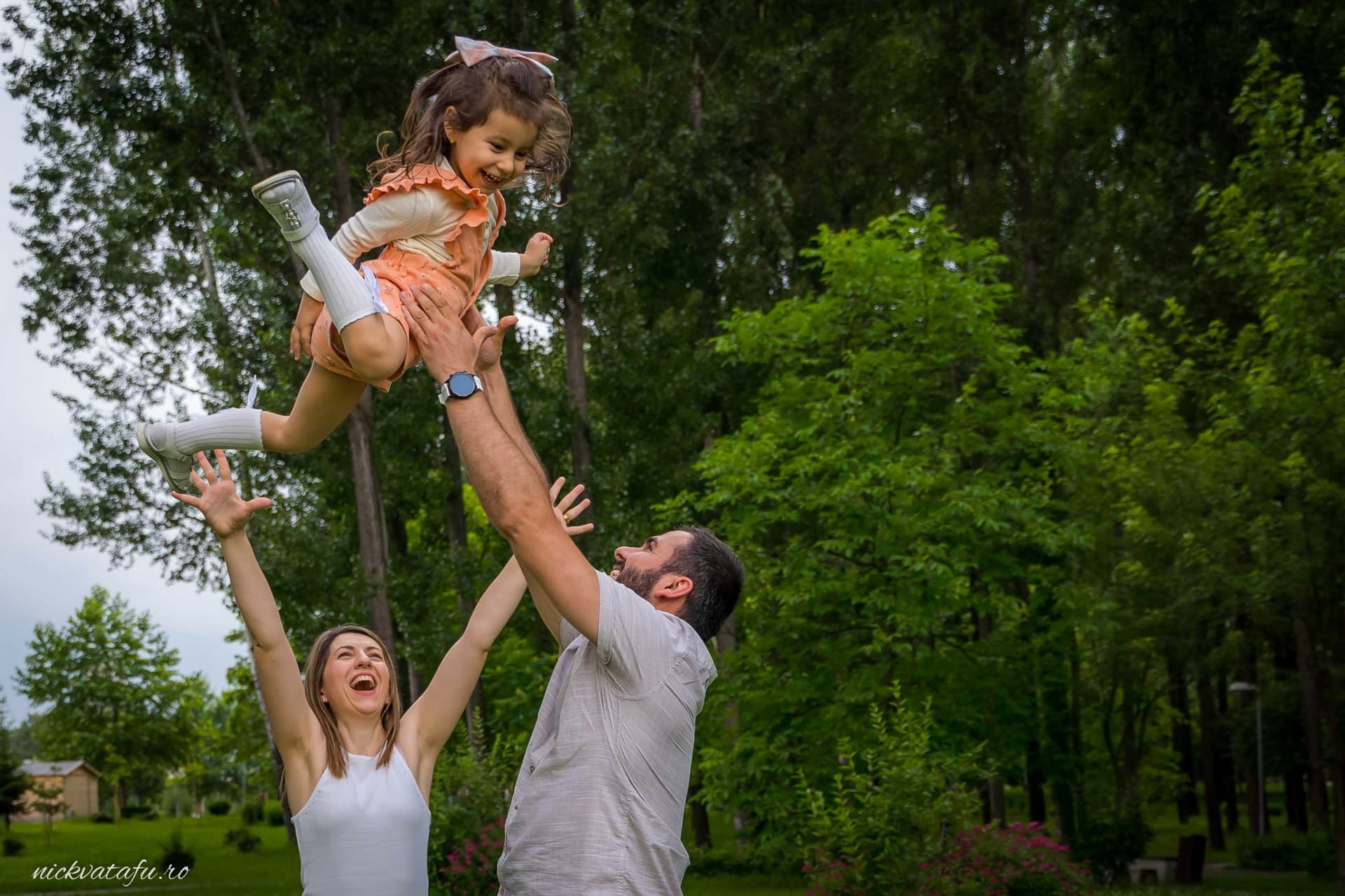 Bărbat și femeie zâmbind, ridicând o fetiță în aer, într-un cadru natural plin de verdeață. Emoție și bucurie de familie.