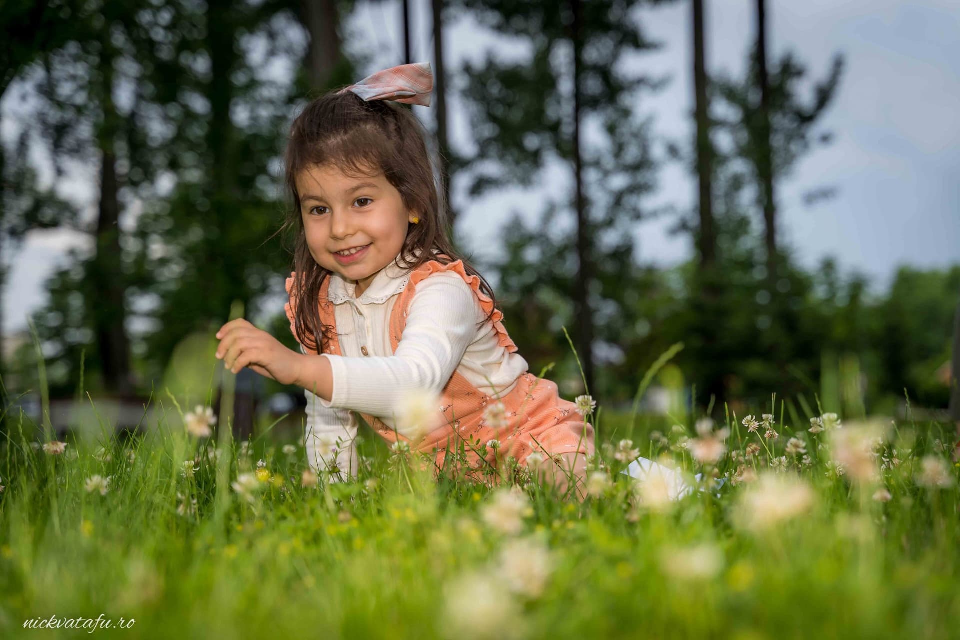 Fetiță zâmbitoare, jucându-se pe iarba verde, înconjurată de flori, evocând bucuria unui botez în natură.